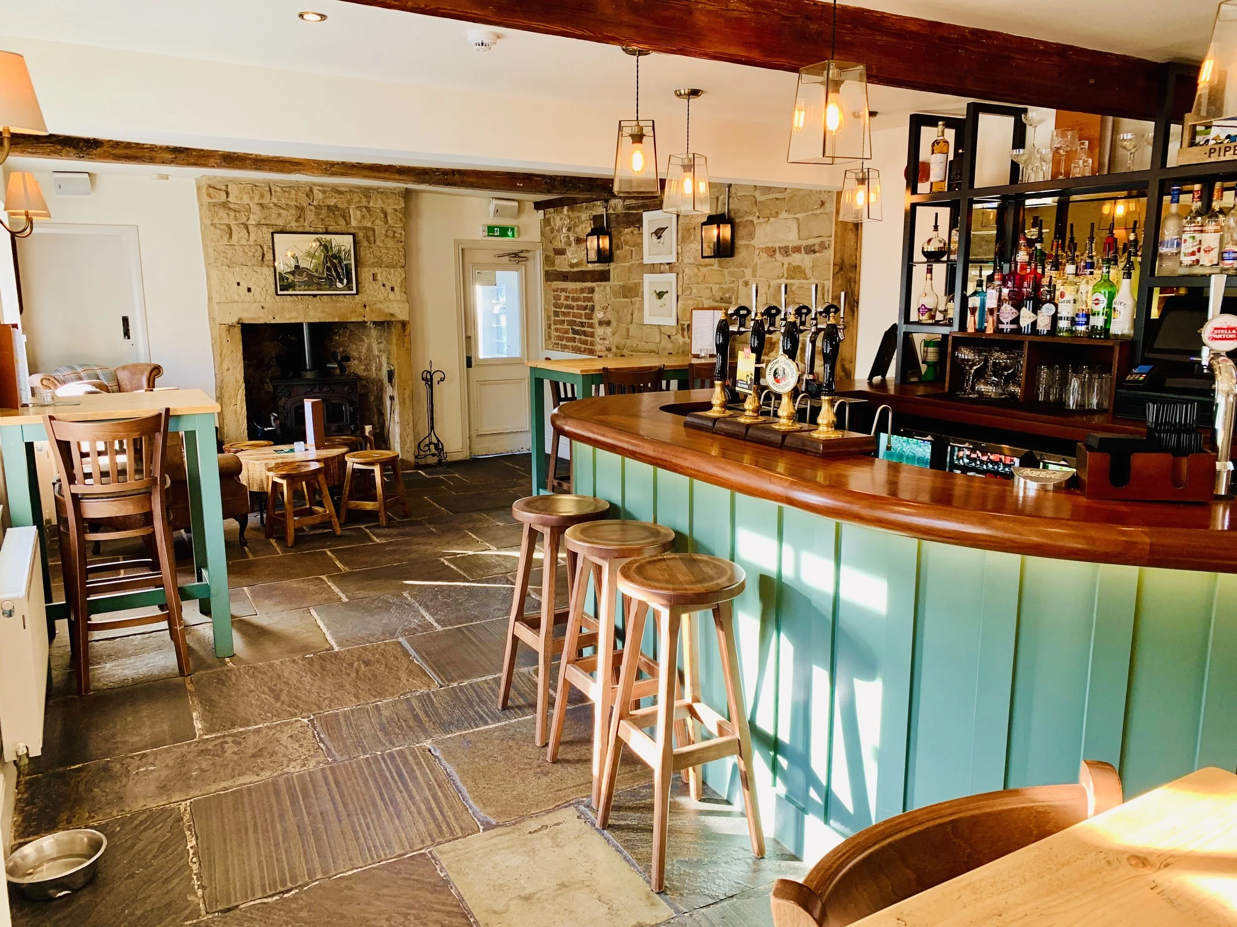 Interior of a cozy pub or restaurant with a curved wooden bar, barstools, a stone fireplace, and various seating areas with tables and chairs.