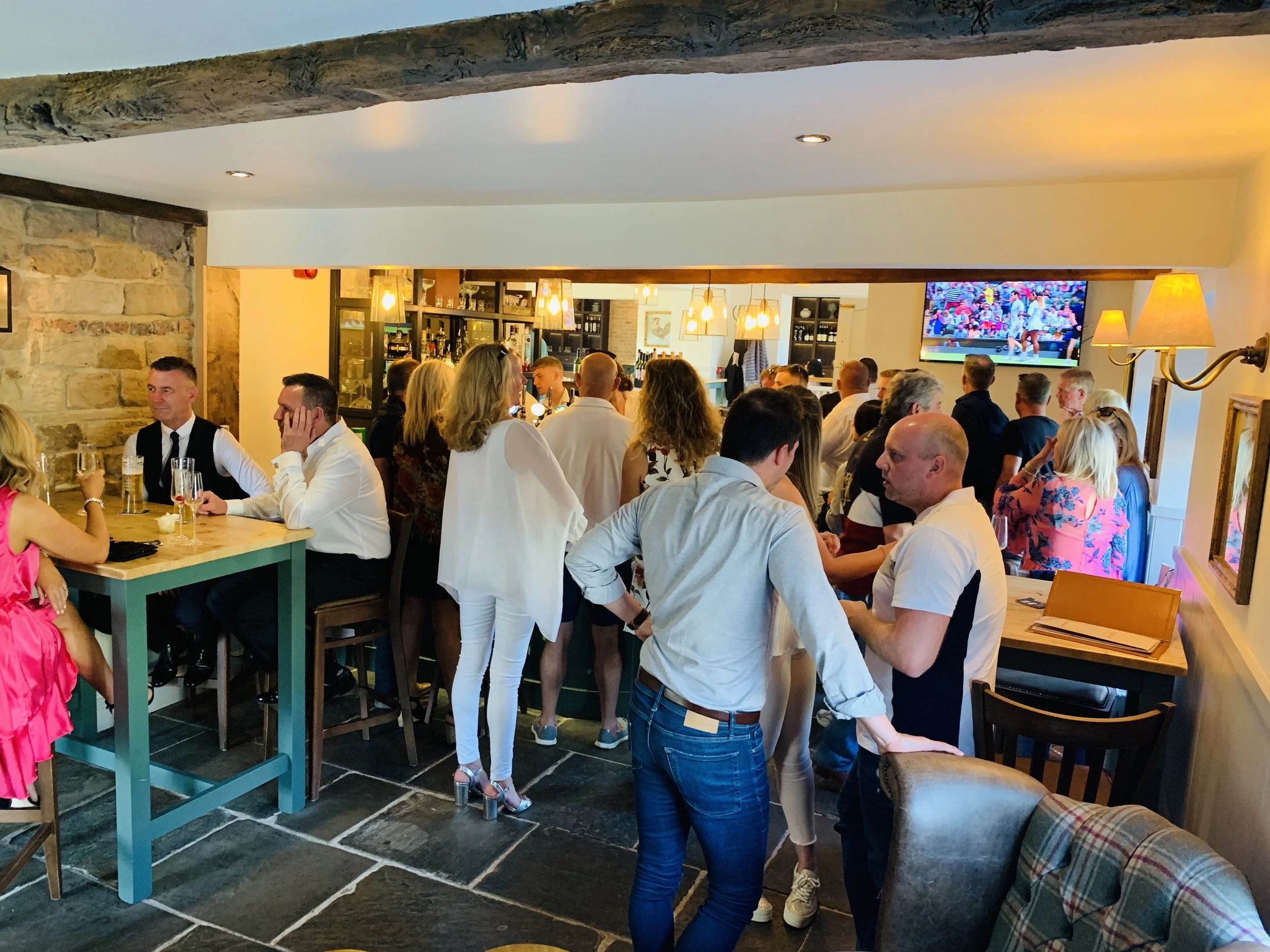 People socializing and watching a sports game on television in a cozy restaurant or pub with stone walls, warm lighting, and casual attire.