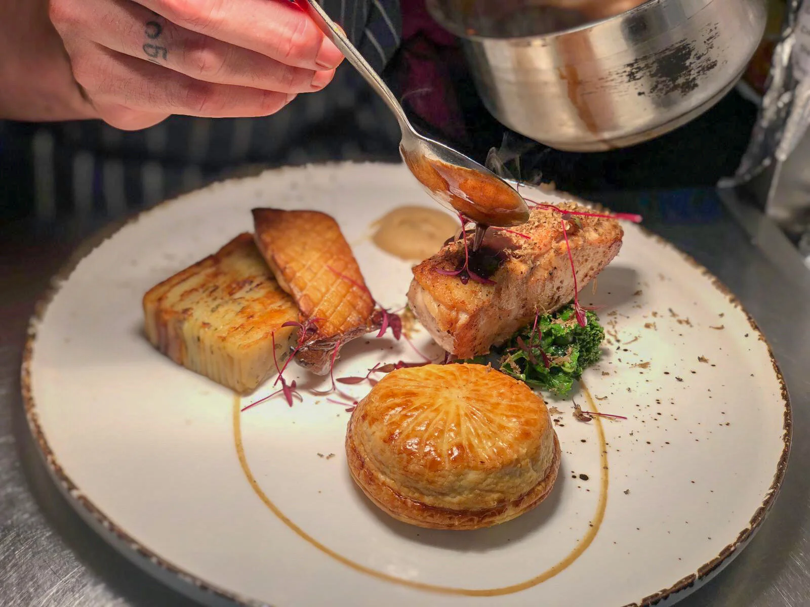 A plated gourmet dish featuring a seared fish fillet with a grilled side, a toasted round pastry, and a colorful vegetable garnish, with a person spooning a sauce onto the fish.