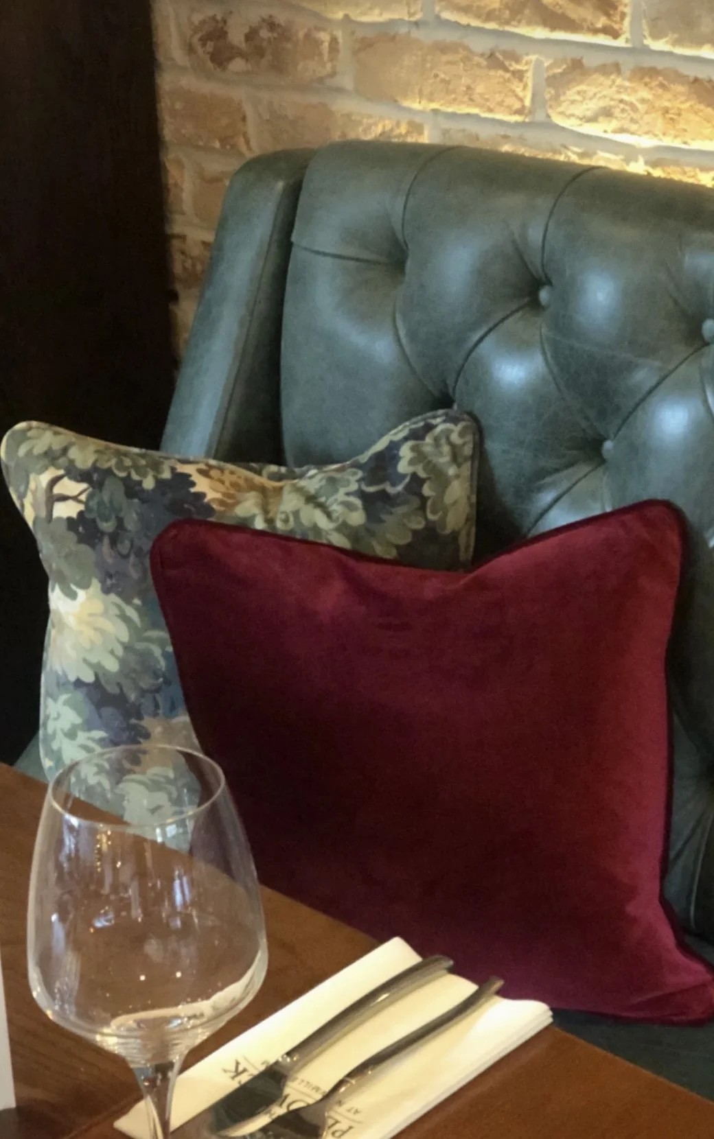 A leather tufted headboard with two decorative pillows, one floral and one solid red, on a bed with a wine glass and utensils on a wooden table beside it.
