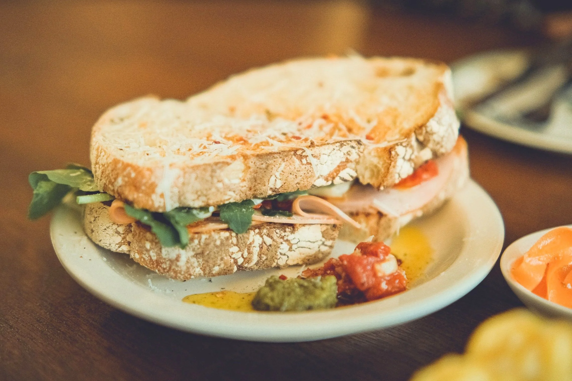 Close-up of a sandwich on a white plate with green and red toppings, accompanied by small dishes of sauce or dips.