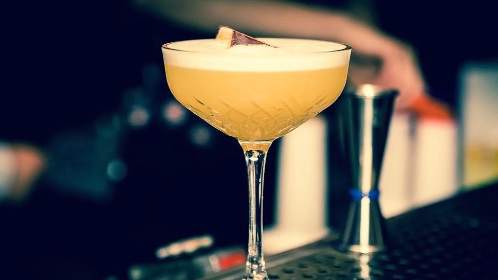 A yellow cocktail with a slice of fruit garnish in a cocktail glass on a bar counter.