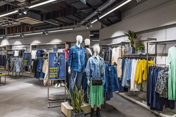 A vintage silver ice cream truck with the sign 'The Little Cafe' on its side, parked on a street near a hedge and a brick wall, with the rear loaded with ice cream freezers.