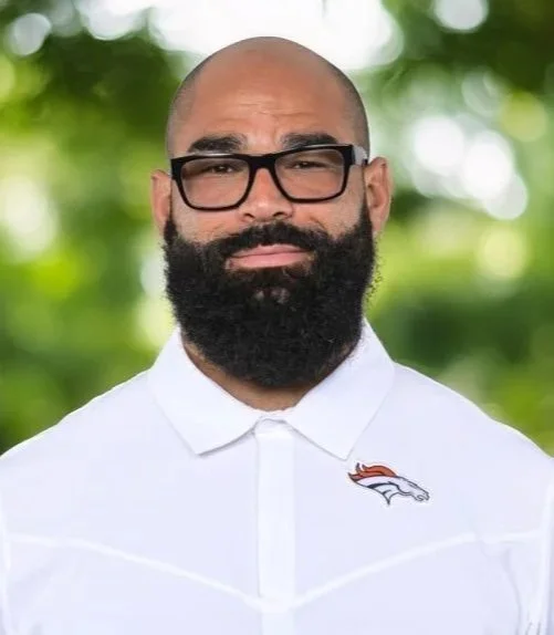 A man with glasses and a beard, wearing a white polo shirt with Denver Broncos logo, outdoors with blurred green trees in background.