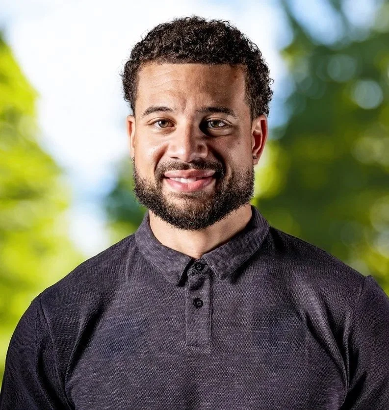 A man with curly hair and a beard, smiling, wearing a dark polo shirt, outdoors with blurred green trees in the background.