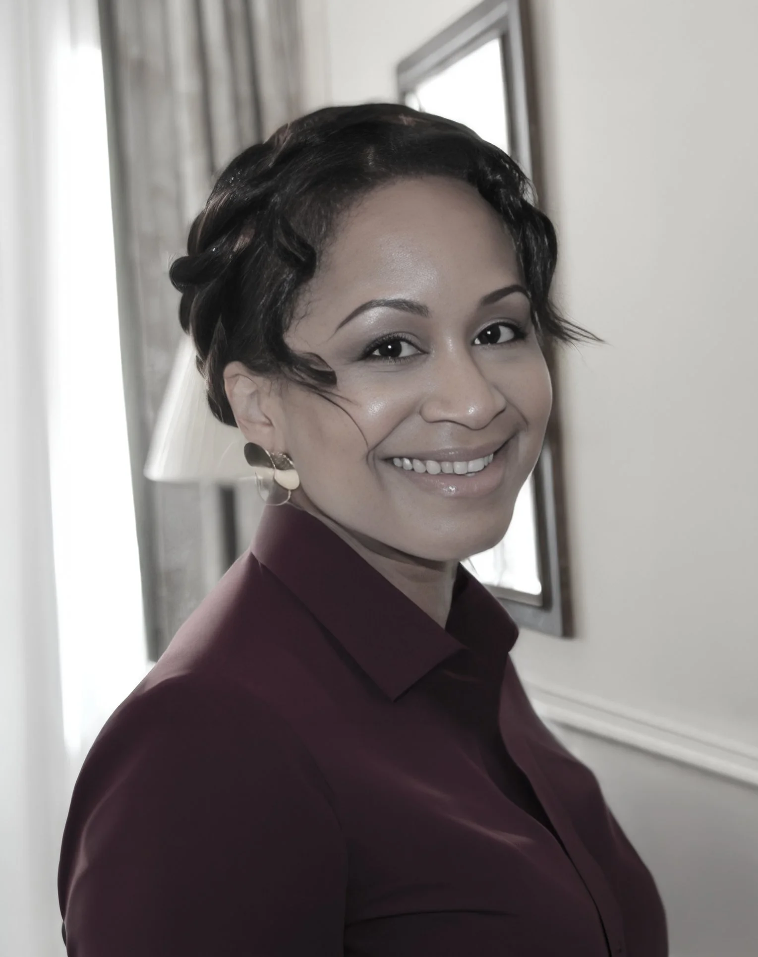 Portrait of a woman with brunette wavy hair, smiling, wearing a burgandy-colored blouse against a light background.