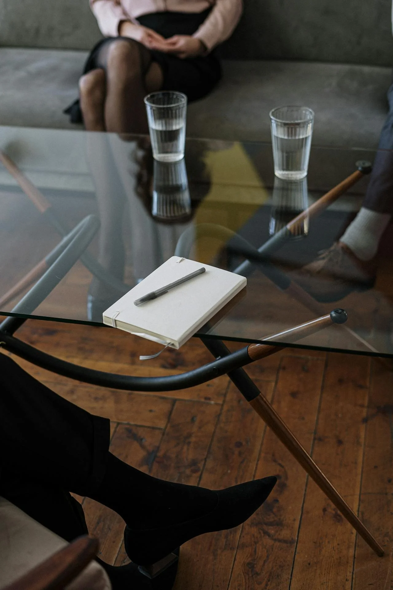 Psychologist session supporting two clients. close up of glass table with notebook glasses of water on it, and three people sitting around.