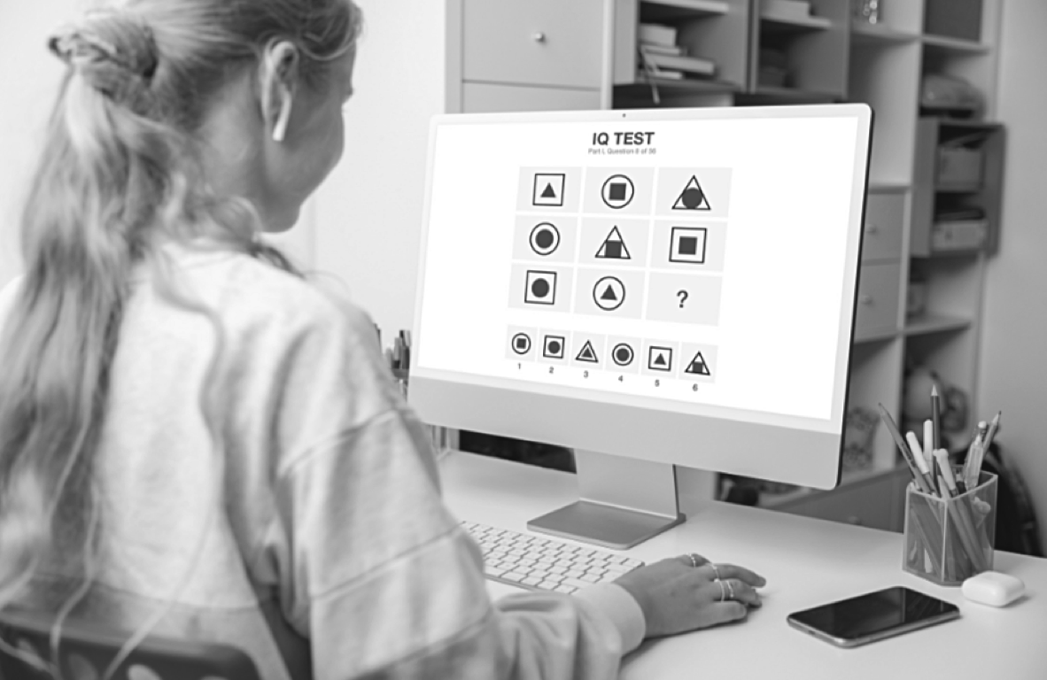 A person with long hair, wearing earphones, seated at a desk, is taking an IQ test on a computer. The test screen shows a series of geometric shape puzzles, with multiple-choice answers below.