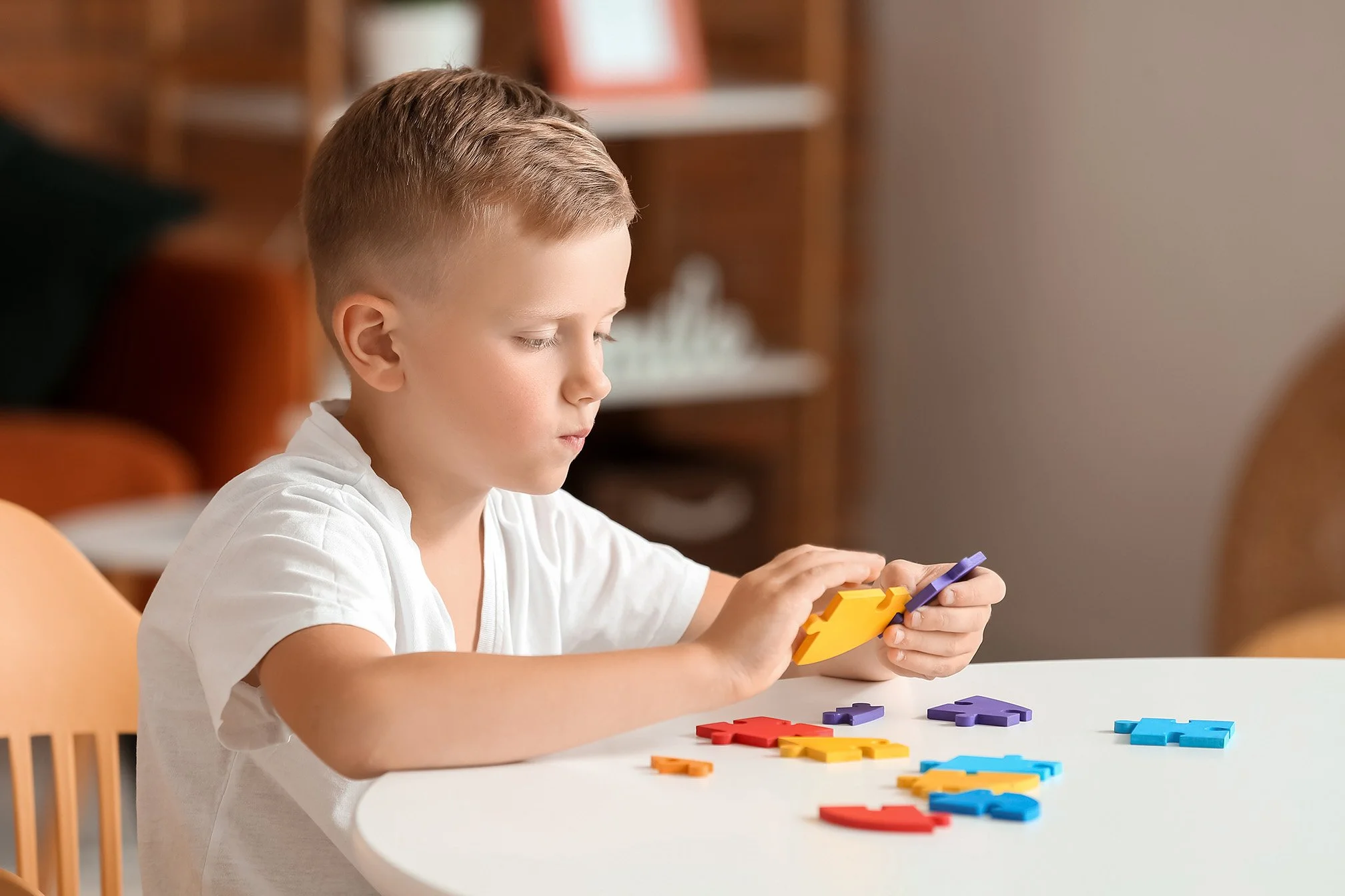 A young boy with short blonde hair sitting at a white table, playing with colorful foam puzzle pieces.