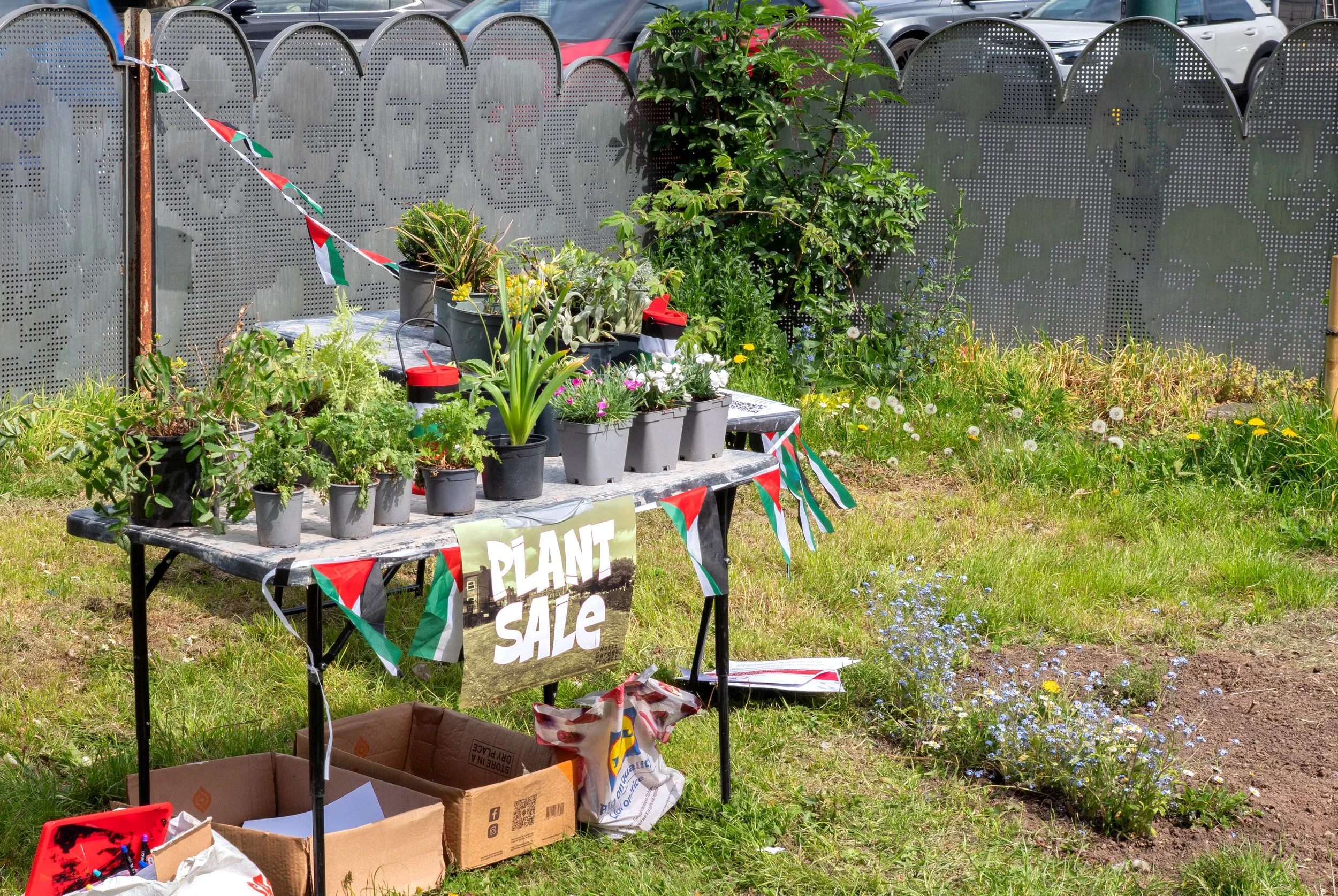 A table in the foreground shows a plant sale and Palestine flag bunting, with flower beds in the background. 