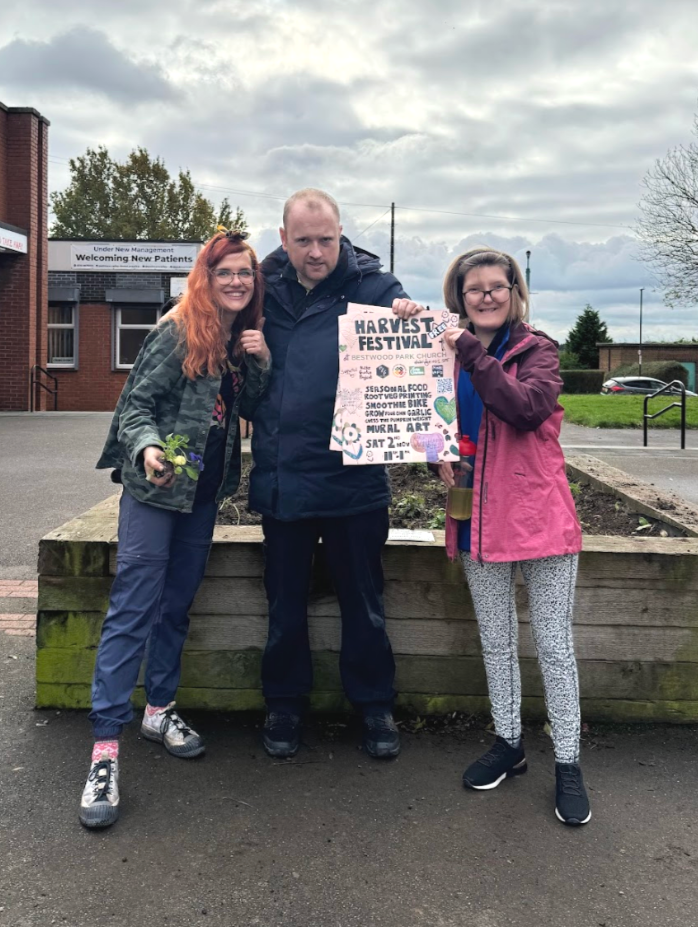 3 poeple stood by a flower bed holding a poster for the harvest festival.