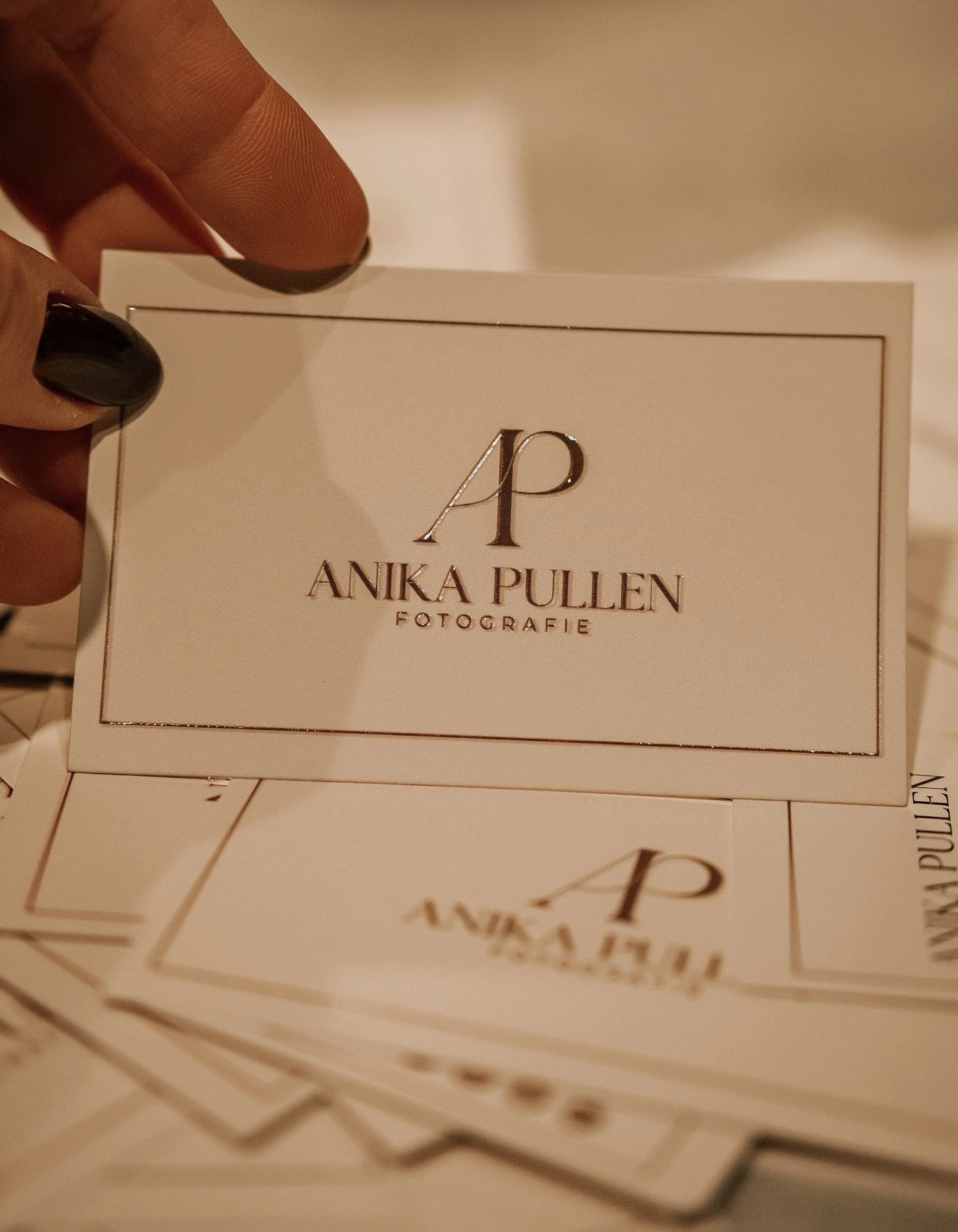 Visitekaartje met tekst "Anika Pullen Fotografie" en initialen "AP", vastgehouden door een hand met zwart gelakte nagels.