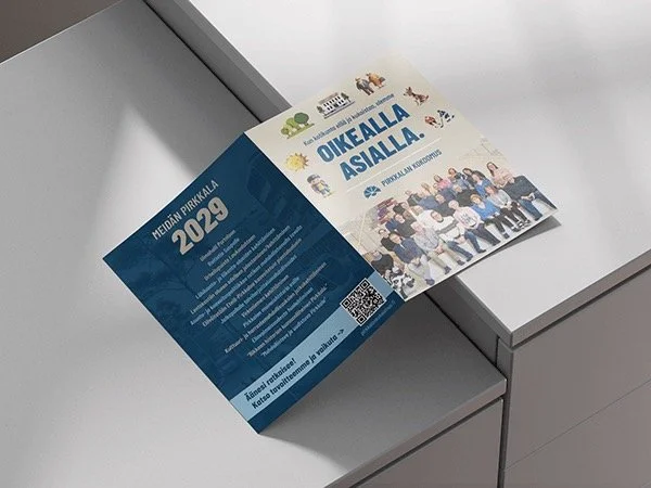 A flyer on a table promoting Meidan Pirkkala 2023 and Oikealla Asialla, with a group photo and colorful illustrations.
