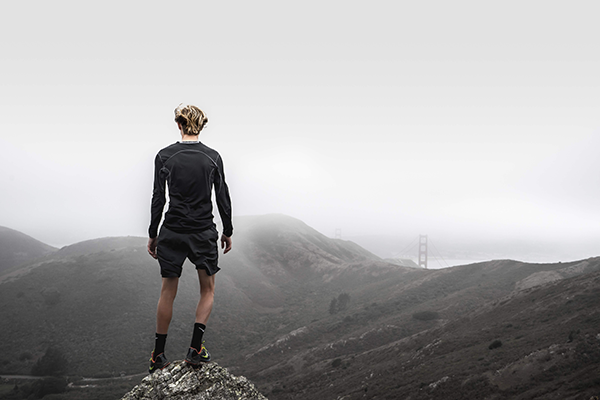 Person standing on a rock overlooking a landscape with hills and a distant bridge.