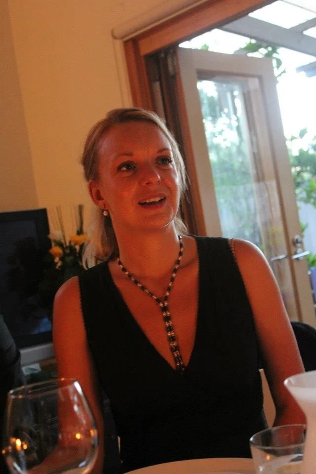 A woman with blonde hair wearing a black sleeveless dress and a black and white beaded necklace, sitting at a dining table indoors with a window showing greenery outside.