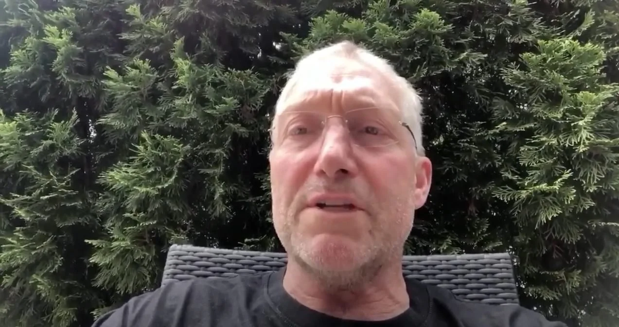 A middle-aged man with white hair and glasses sitting outside in front of a large evergreen tree.