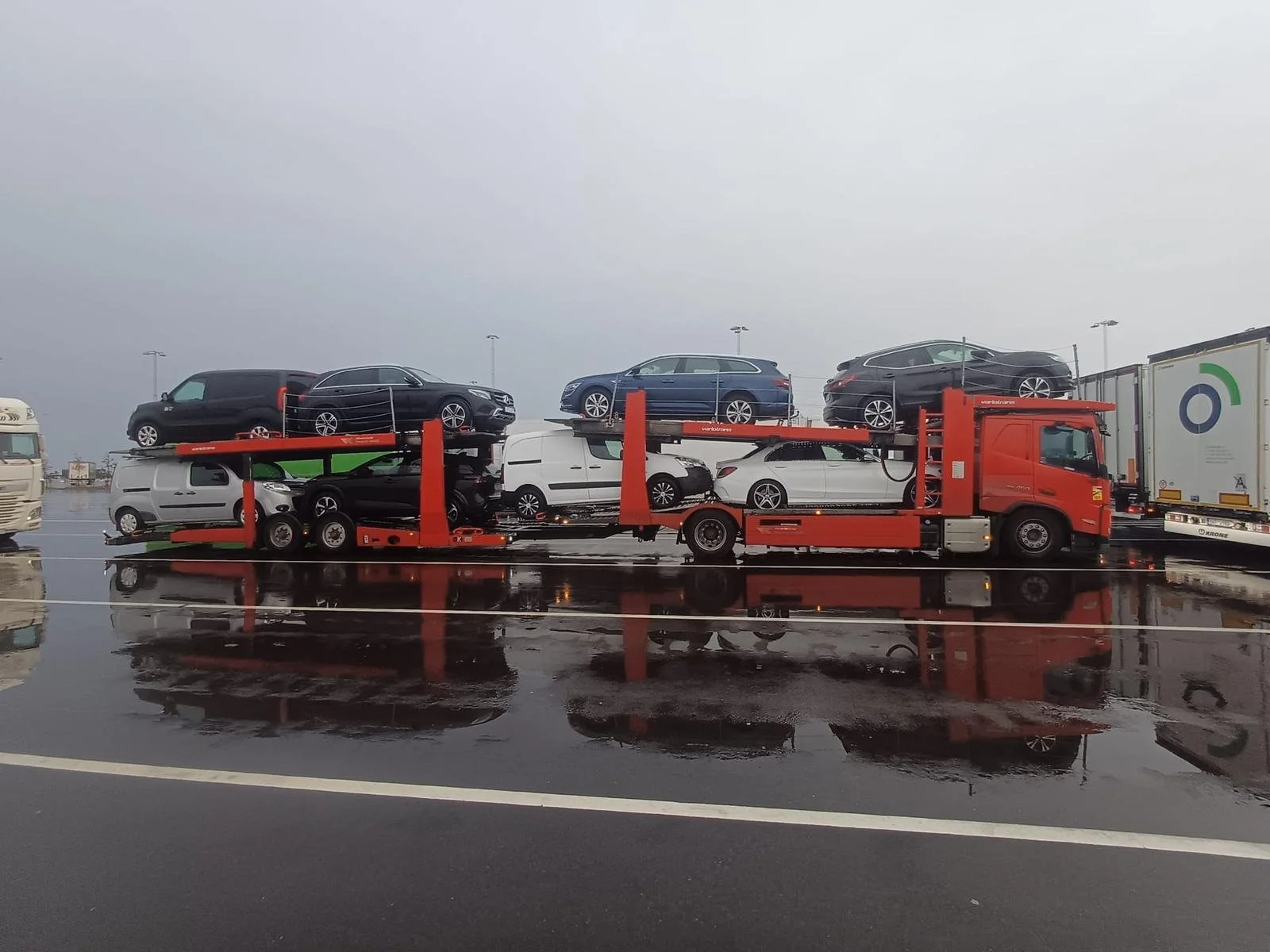 A red car transporter truck parked on a wet surface loaded with multiple cars, including SUVs and sedans, arranged in two levels. Other vehicles, including trucks, are visible in the background.