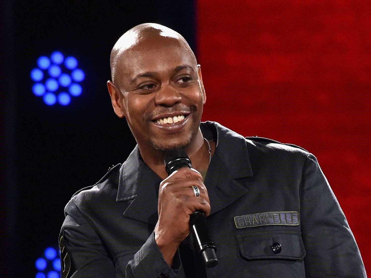 Person holding a microphone and smiling on stage, wearing a dark jacket with name patch "Chappelle."
