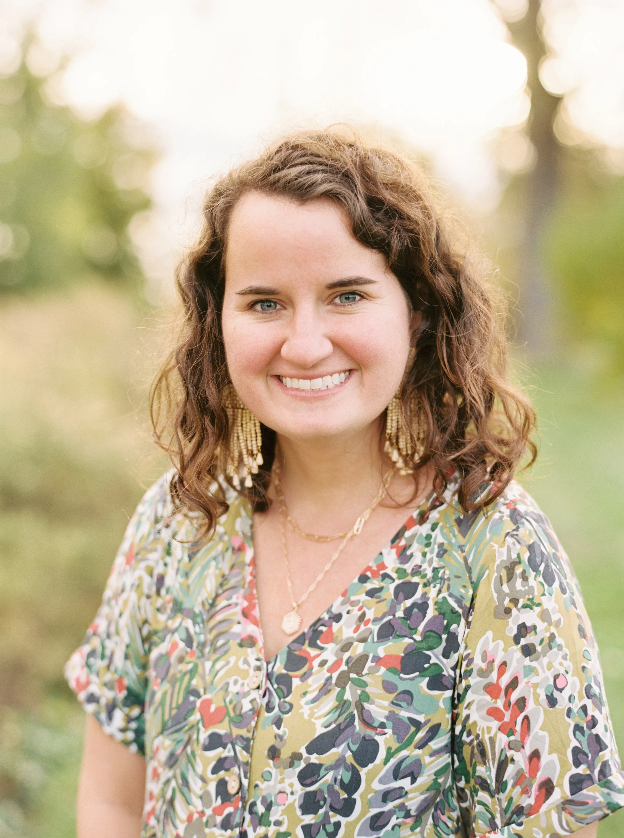 A woman with curly brown hair and blue eyes smiling outdoors in a park or garden, wearing a colorful patterned dress and gold jewelry.
