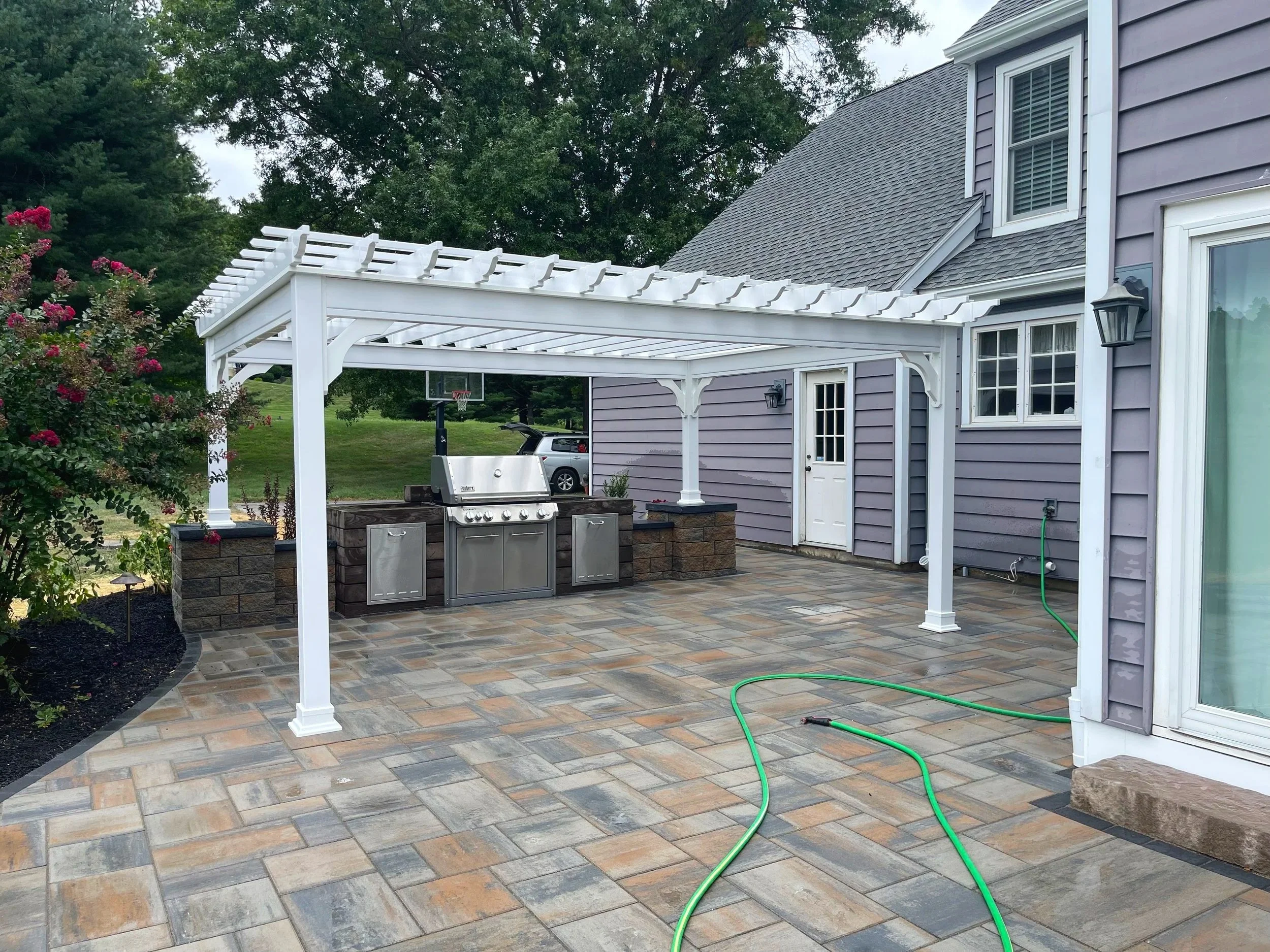 A backyard patio with a white pergola, outdoor grill, potted plants, and a house with purple siding. Green garden hose and parking lot with a car and a basketball hoop in the background.