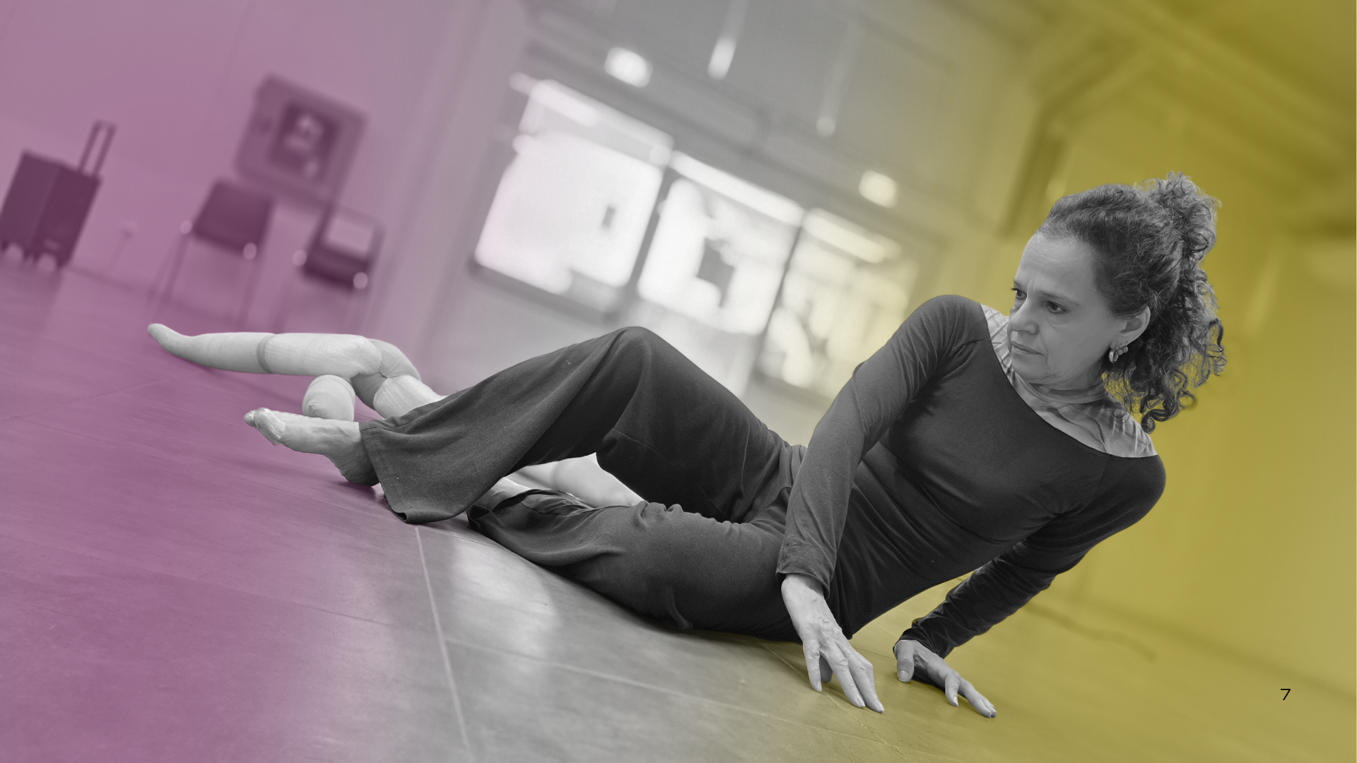 Una donna con capelli ricci e scondizionati, indossa un abbigliamento casual e si trova su un pavimento, in vecchio stile, in una stanza luminosa con grandi finestre. La stanza è colorata aufoni e viola, con alcune sedie e un televisore sullo sfondo.