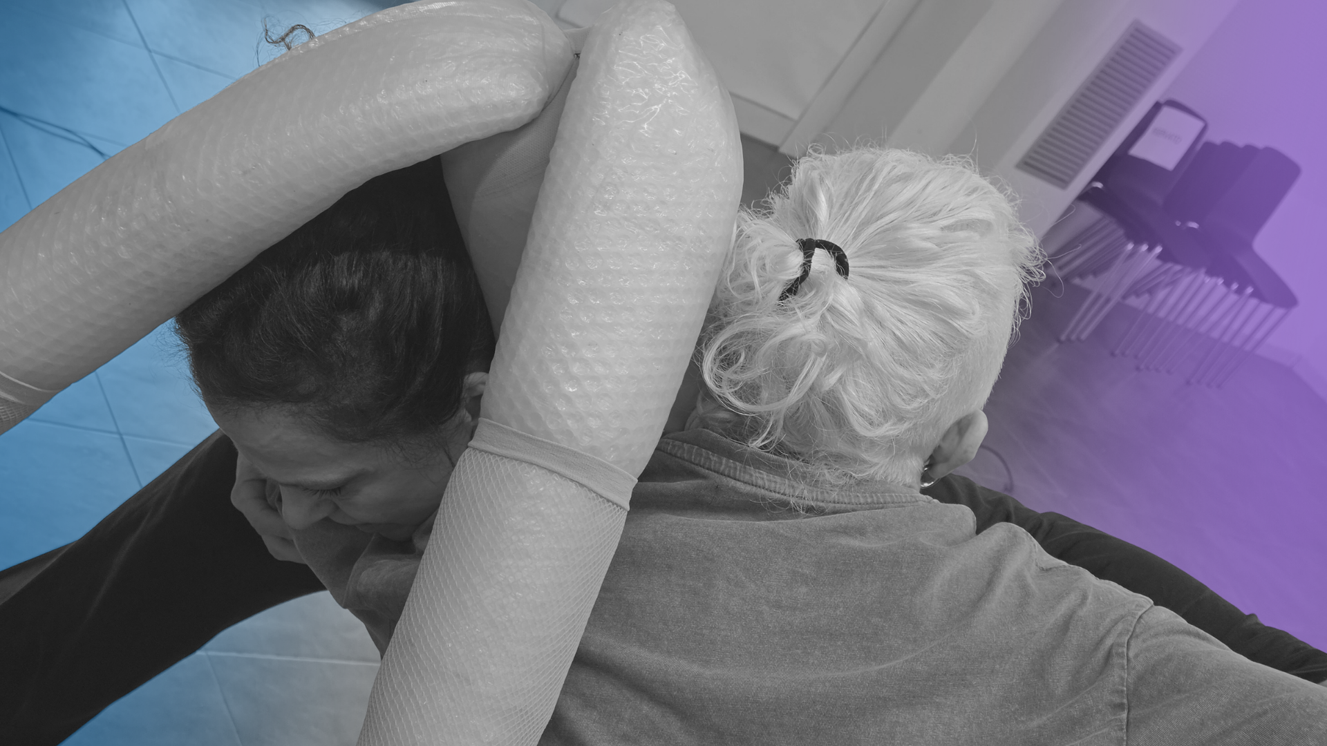 Due persone stanno praticando una dimostrazione di primo soccorso, una delle quali indossa un braccio di protezione con materiale che sembra foam, in un'area interna.