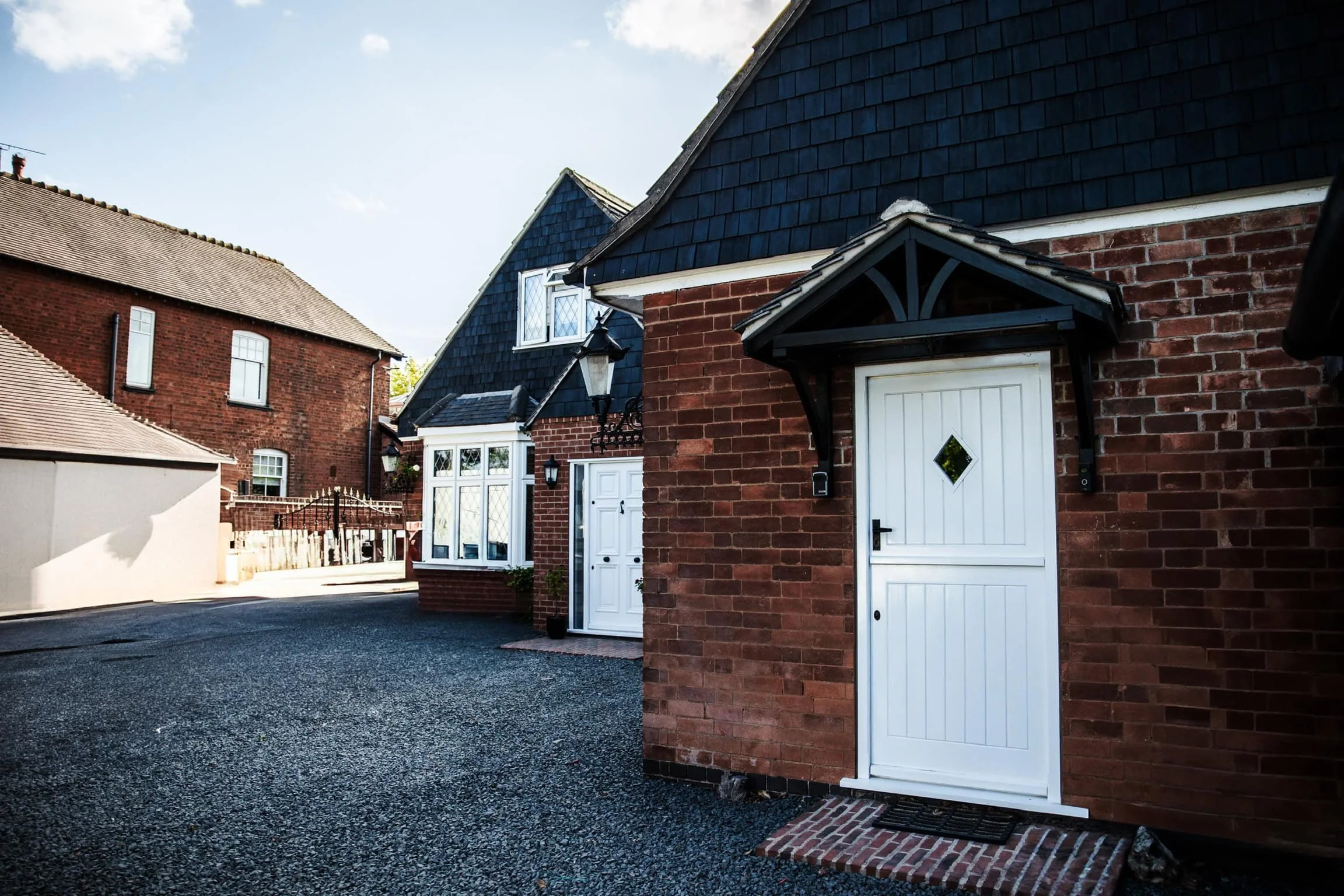 View of a brick house with a white door, black decorative awning, and a small diamond window, situated on a gravel driveway in a neighborhood with other brick houses.