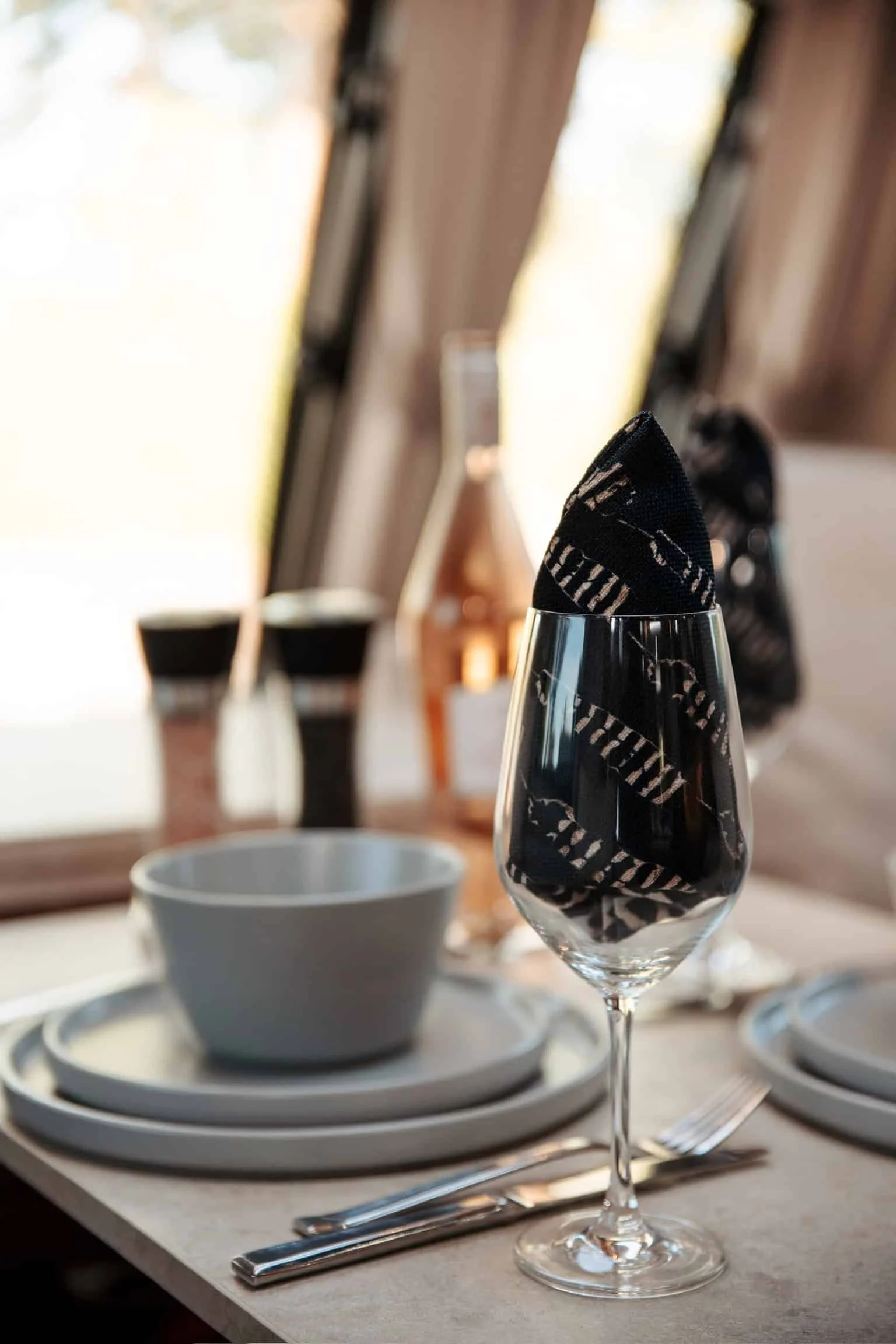 Elegant table setting with a wine glass holding a black cloth napkin, white plates, bowls, and cutlery on a beige tablecloth, with a window in the background.