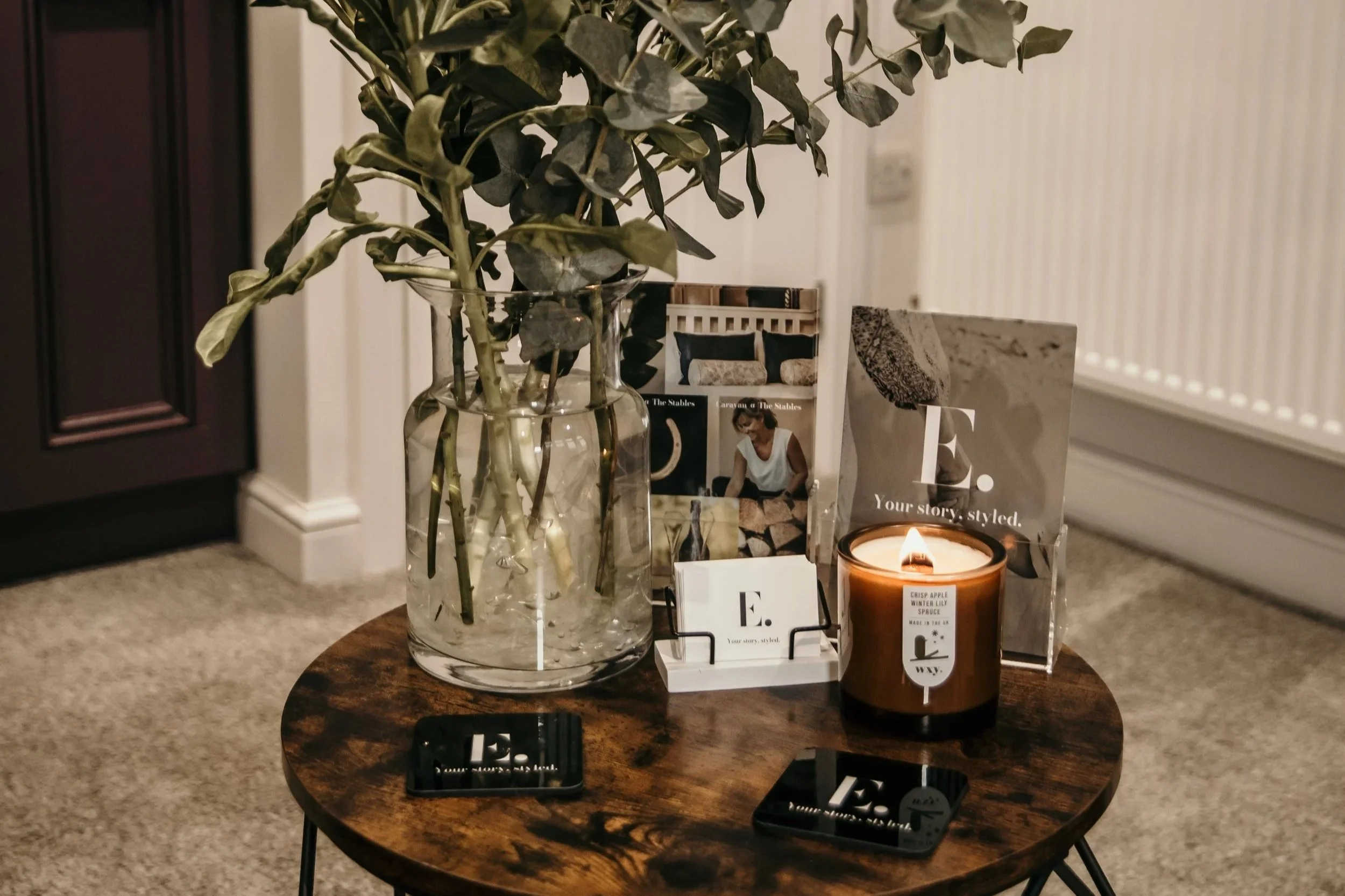 A round wooden table holds a large glass vase with green leafy branches, a lit candle in a brown container, and various promotional cards and pamphlets with the letter 'E' on them, placed on a magazine holder.
