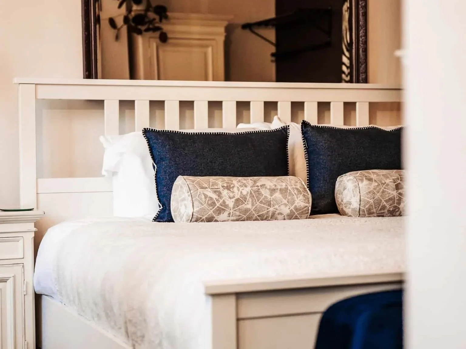 Close-up of a neatly made bed with white bedding, dark blue pillows with decorative stitching, and beige cylindrical bolster pillows with a leaf pattern, all against a white wooden headboard.