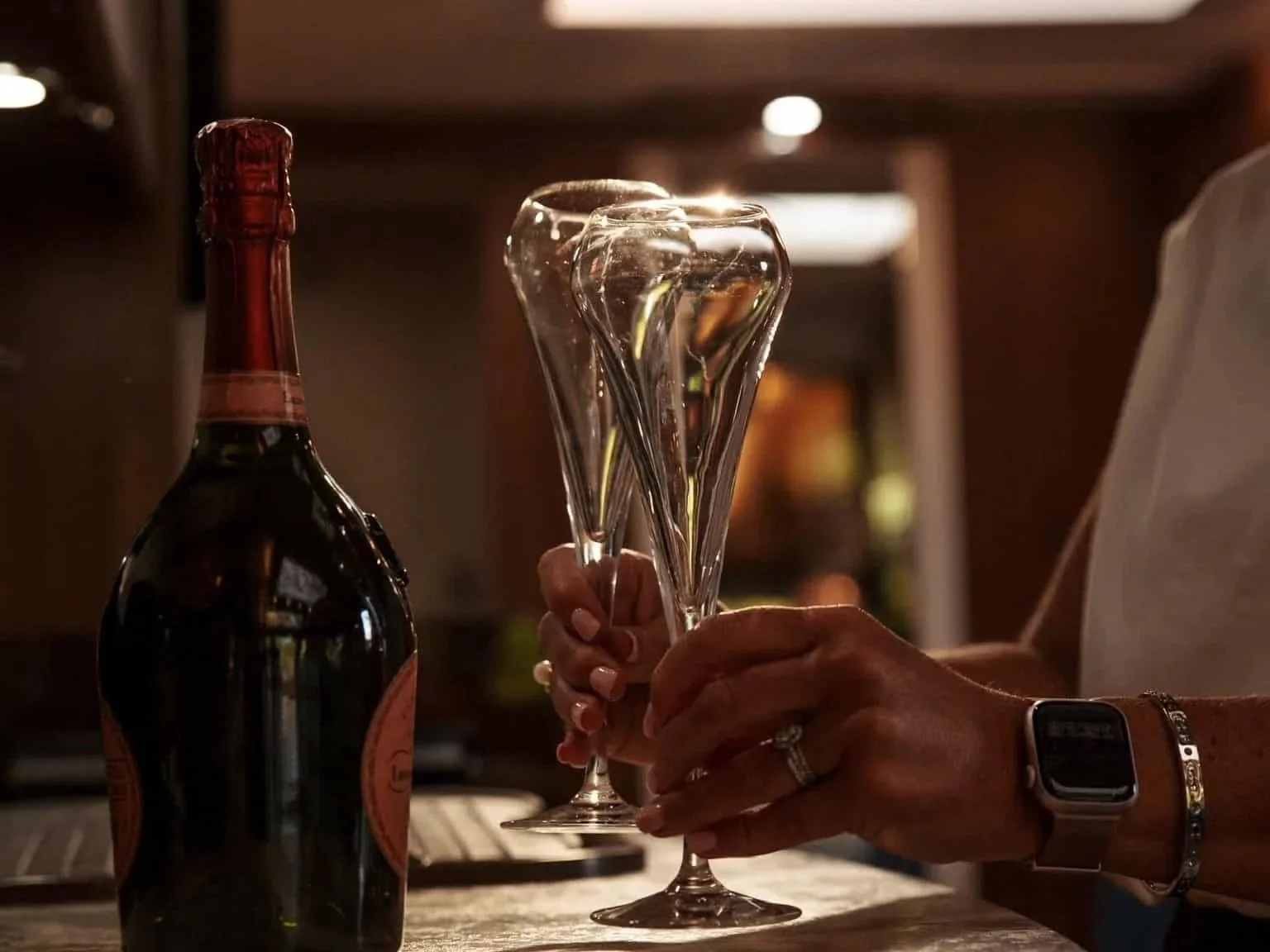 Person holding three empty champagne glasses near a bottle of champagne on a table.