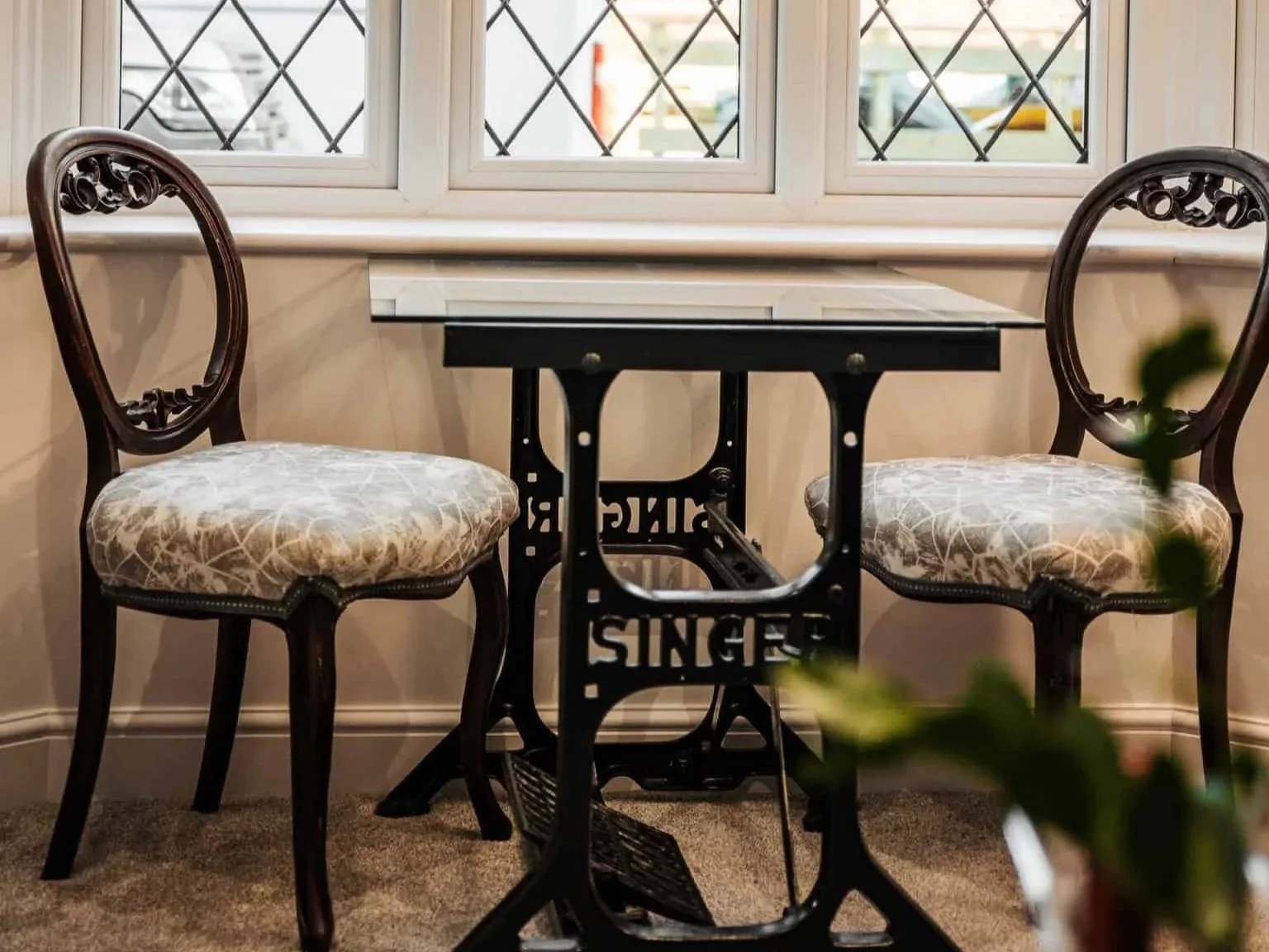 A vintage sewing table with two ornate wooden chairs with cushioned seats, positioned in front of a window with diamond-shaped panes. The table has the words 'SINGERS' on its base.