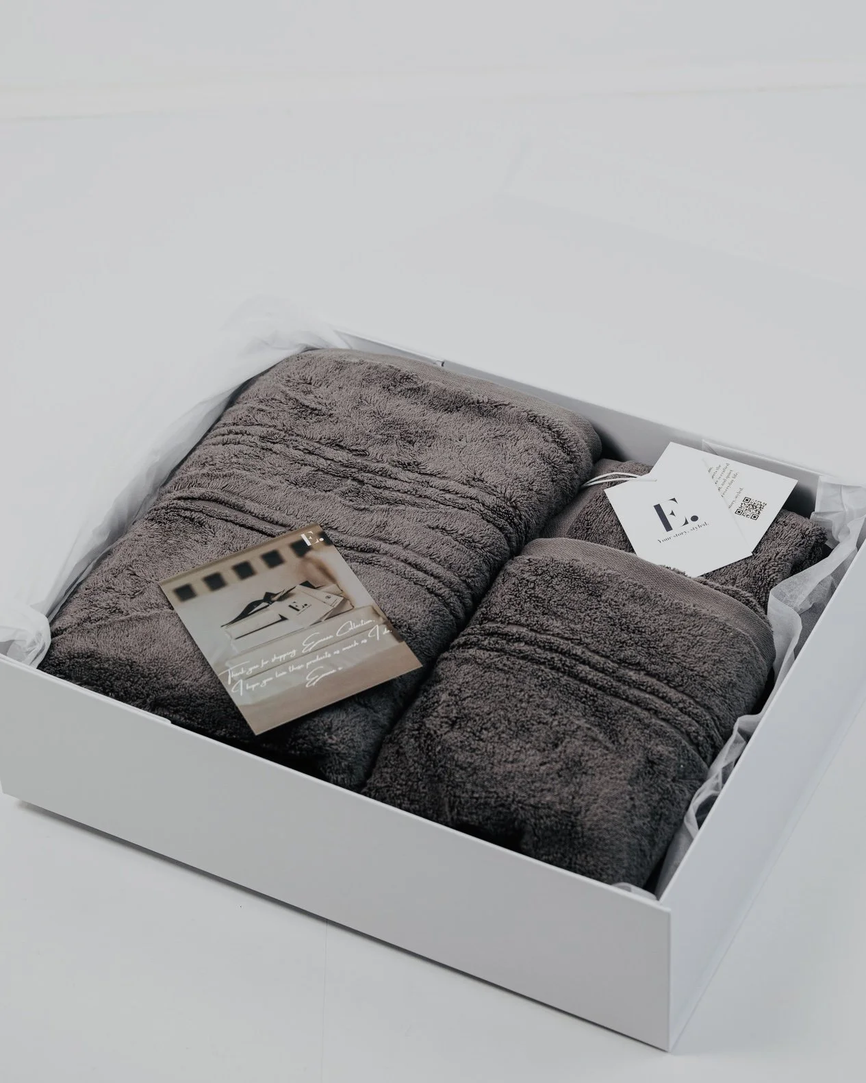 A white box containing two folded dark gray towels, with a small business card and a product tag inside.