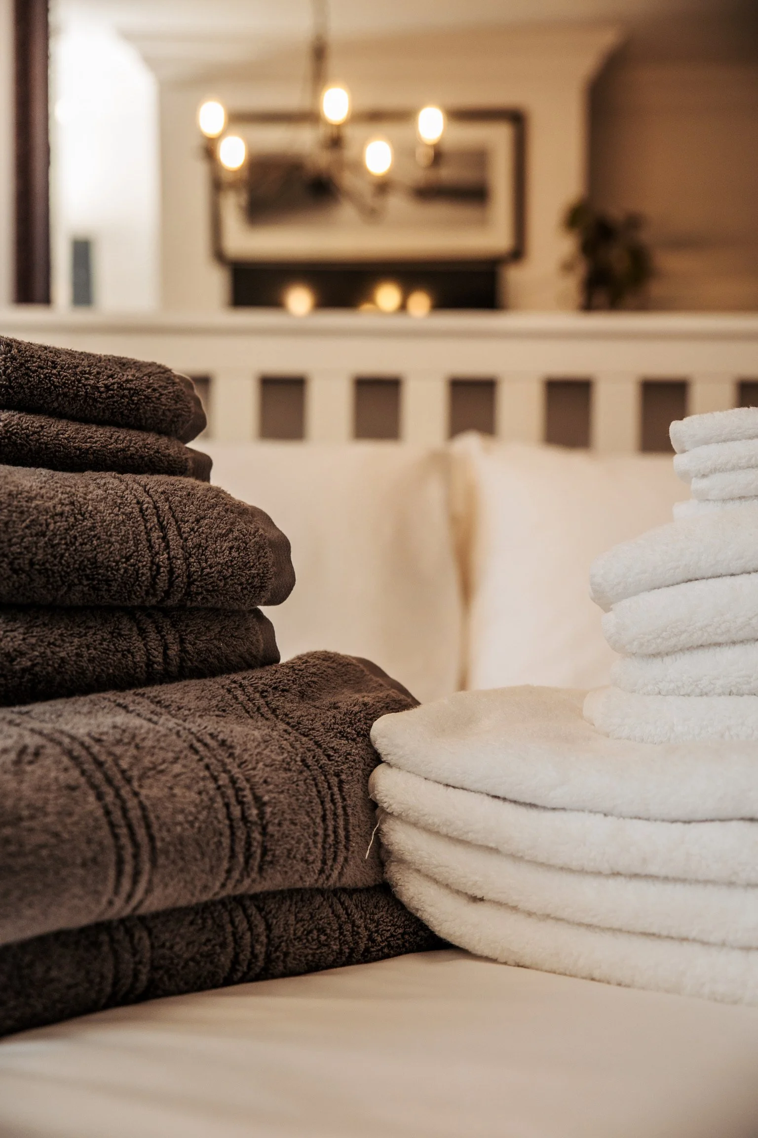 Stacks of folded towels, with dark brown towels on the left and white towels on the right, on a bed with a headboard in the background.