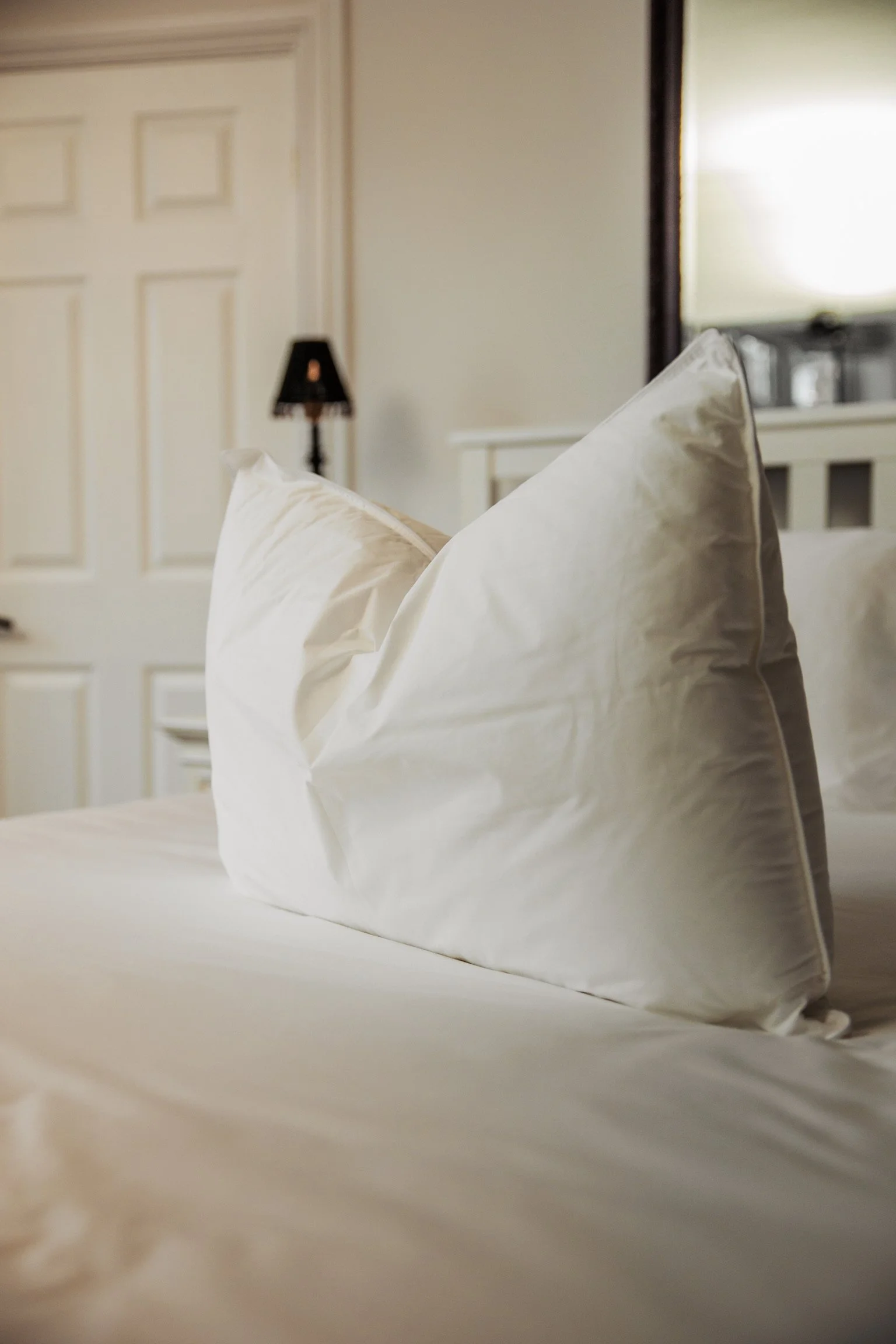 Close-up of a white pillow on a made bed in a cozy bedroom with a lamp and a mirror in the background.