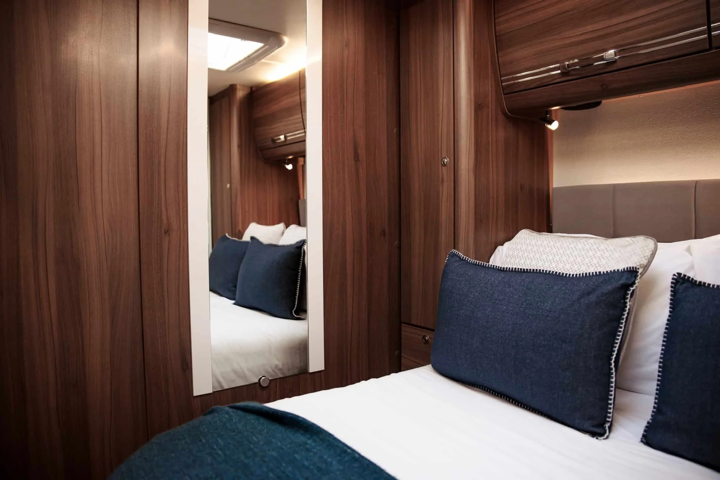 A bedroom with wood-paneled walls, a bed with white bedding, navy blue pillows, and a mirror reflecting part of the room.