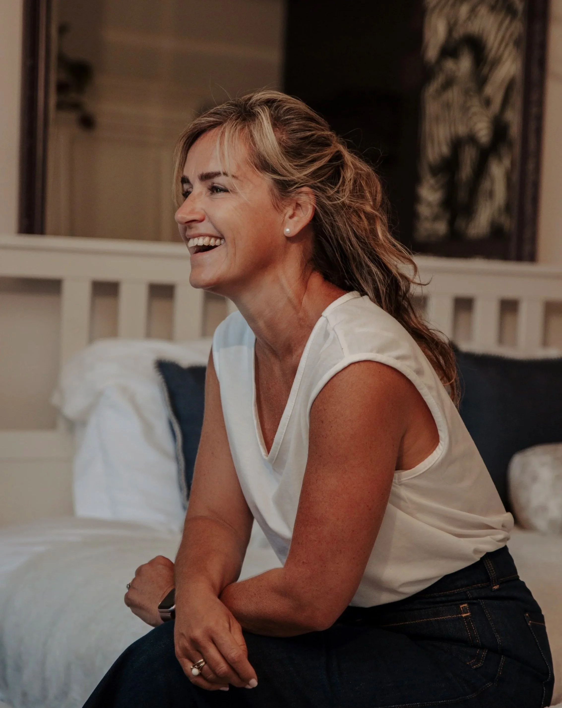 Smiling woman with light brown hair sitting on a bed in a bedroom.