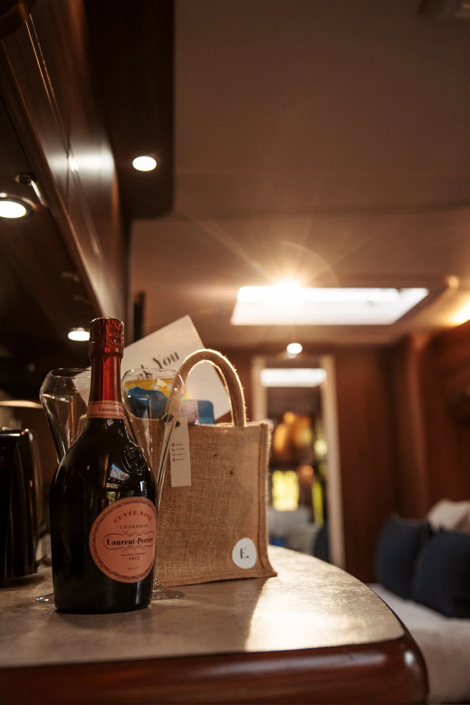 A bottle of Laurent-Perrier Champagne, two empty wine glasses, and a small gift bag on a wooden table in a warmly lit room.