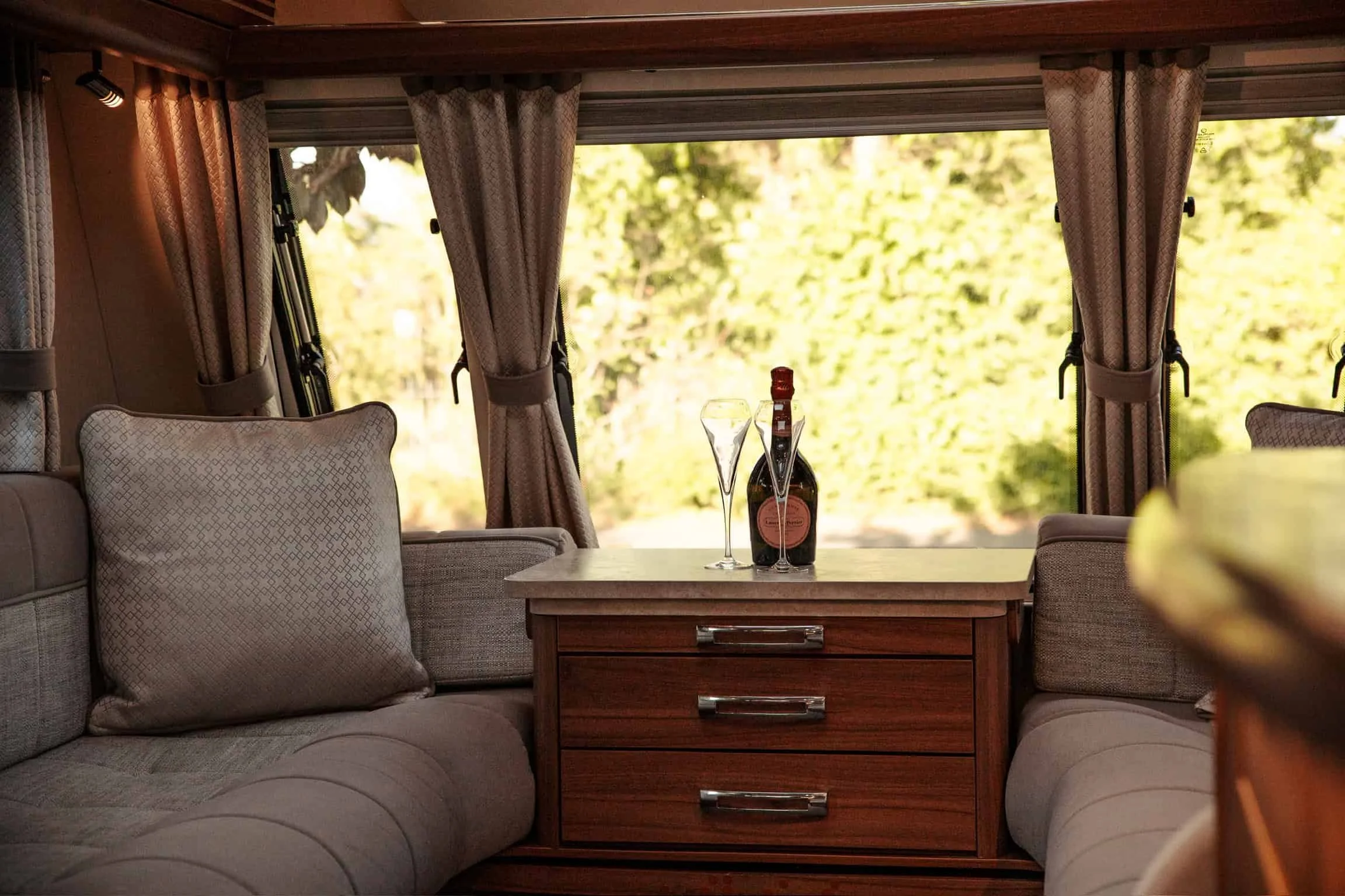 Inside a cozy RV, a table holds two glasses of champagne and a bottle of wine. The scene is framed by beige curtains and cushioned seating, with a view of greenery outside the window.