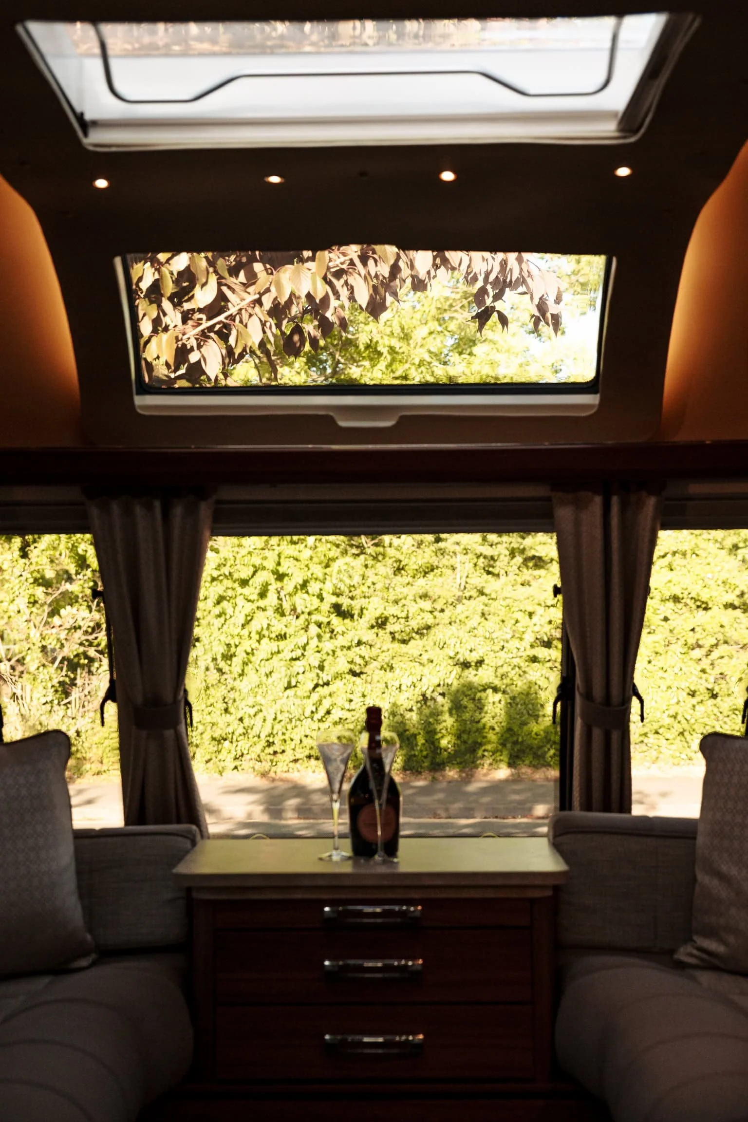A cozy sitting area inside a bus or camper with a small table, bottle of wine, two glasses, and curtains framing a view of green bushes outside. A sunroof or skylight is above, allowing natural light into the space.
