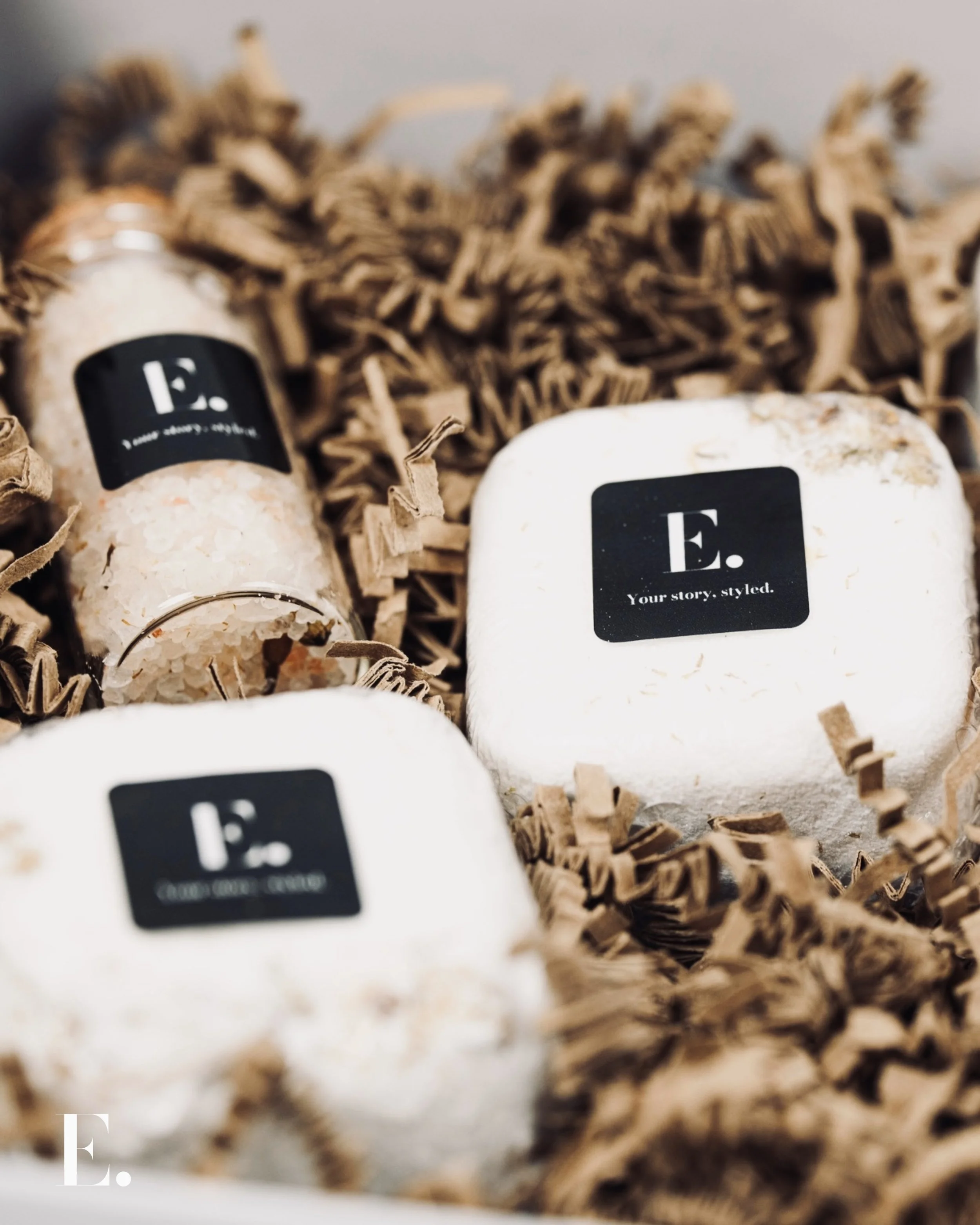 Soap bars and bath salts in a gift box with shredded paper filler, black labels on the products with white text, one label reads 'E. Your story, styled.'