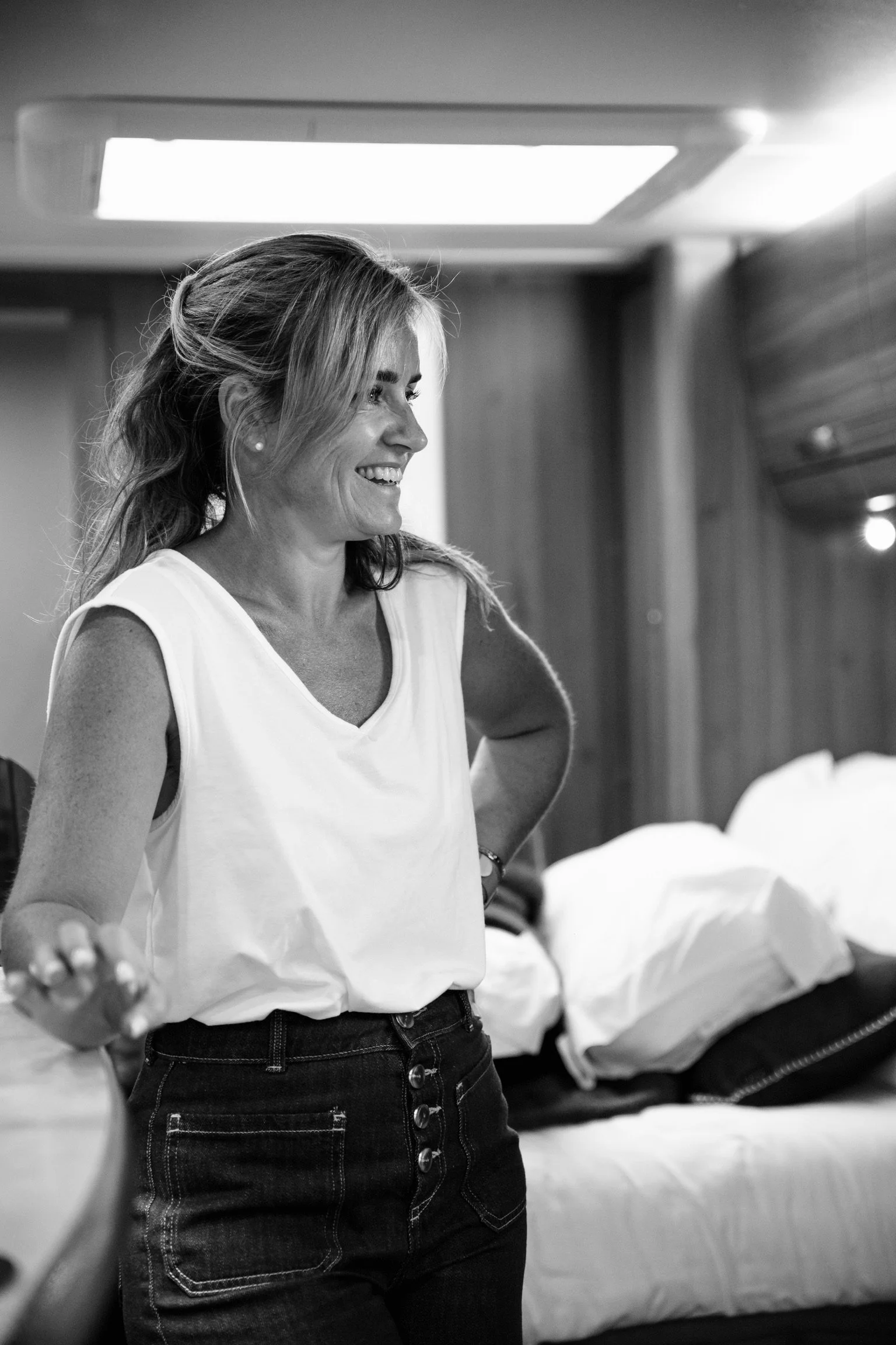 A smiling woman with shoulder-length hair wearing a sleeveless white top and high-waisted jeans, standing in a room with a bed and wooden panel walls, engaging in conversation.