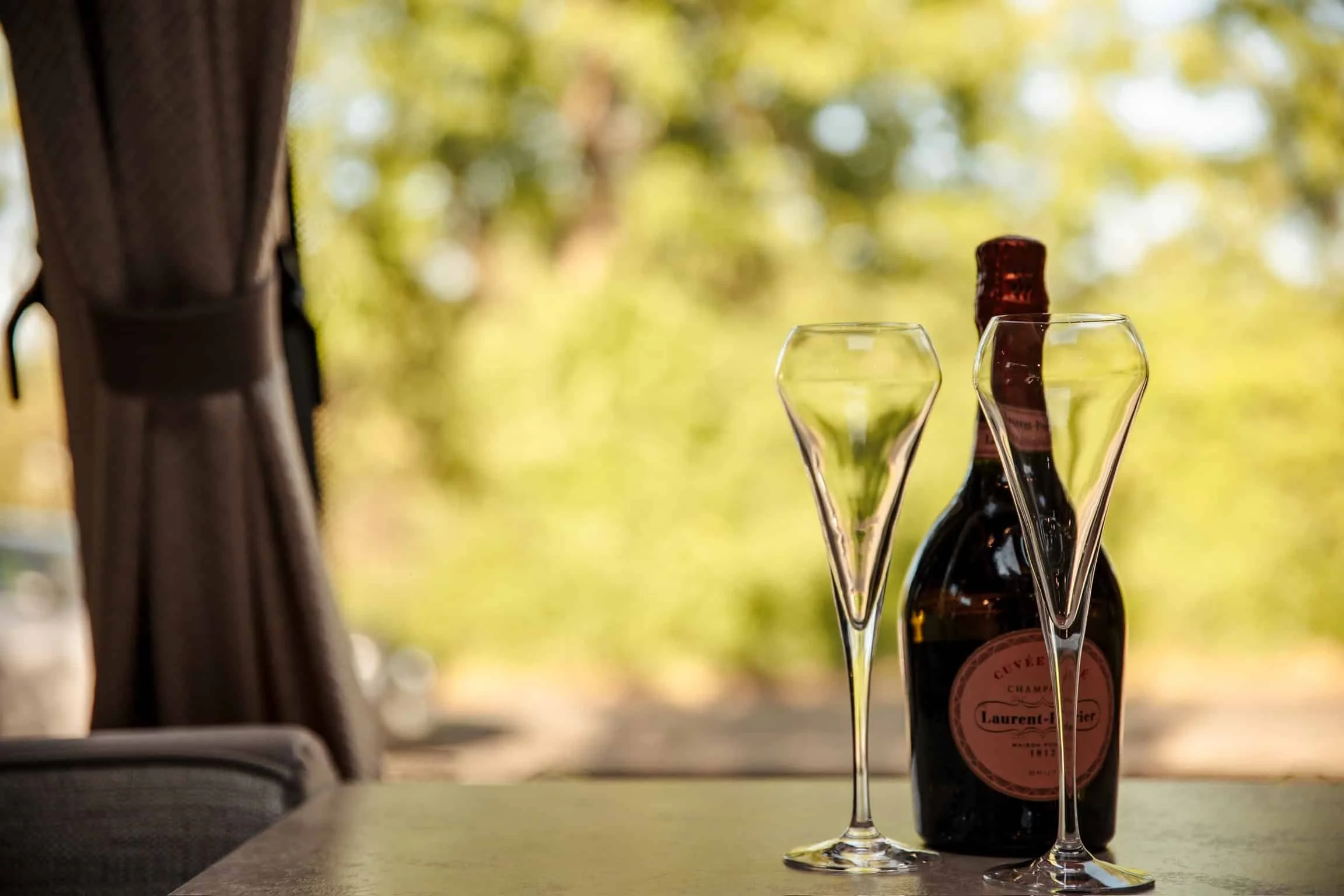 A bottle of Laurent-Perrier champagne and two empty champagne glasses on a table, with a blurred outdoor background of trees and sunlight.