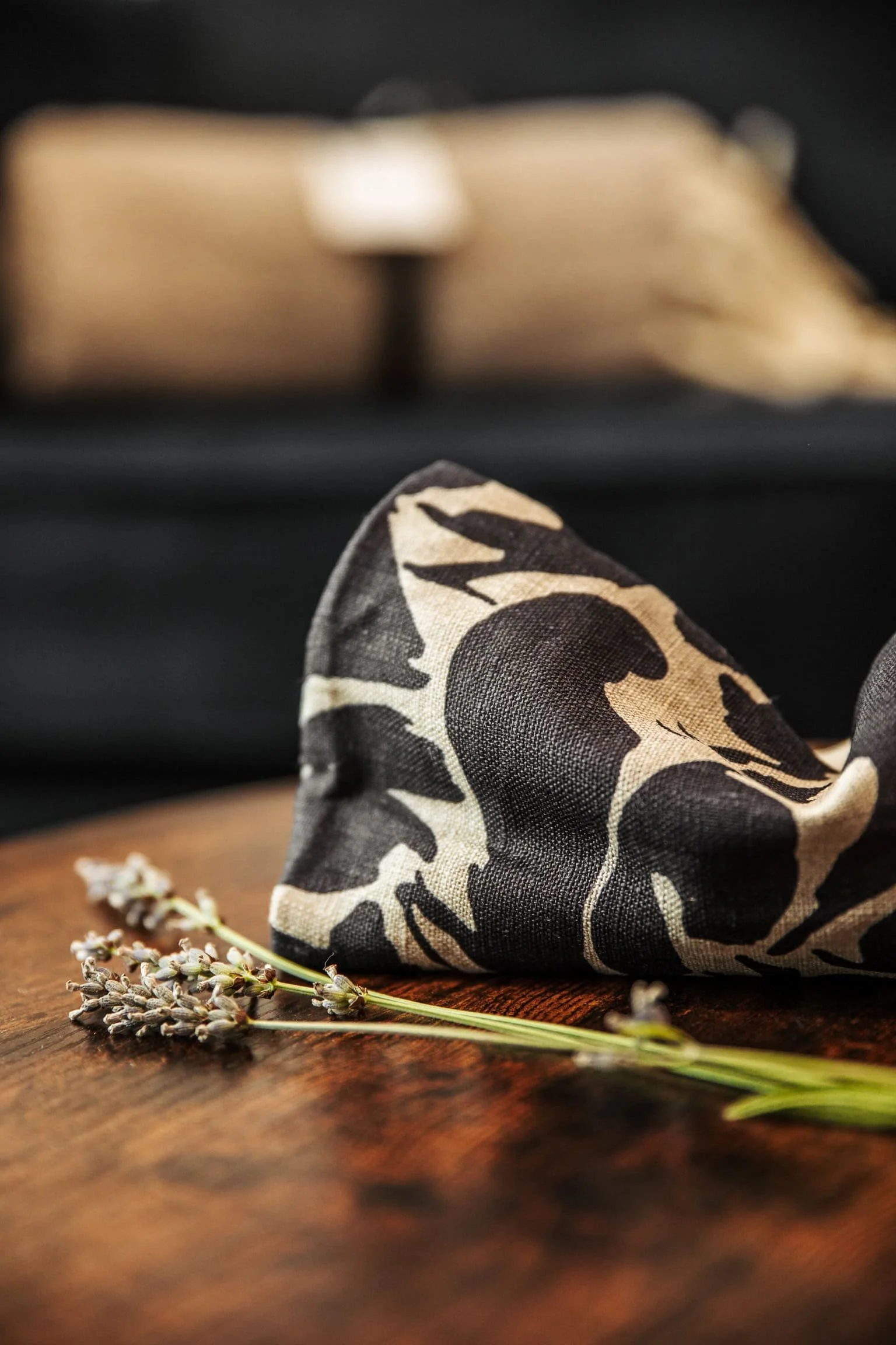 Close-up of a black and beige patterned cloth napkin on a wooden surface with a sprig of lavender flowers in front, and a blurred open book in the background.