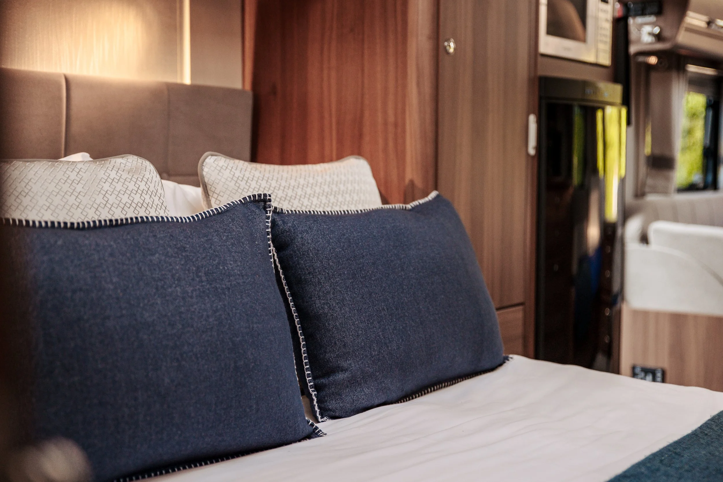 A cozy bedroom with navy blue pillows on a bed, beige and white pillows behind, and wooden walls with a built-in cabinet and a TV in a small living space.