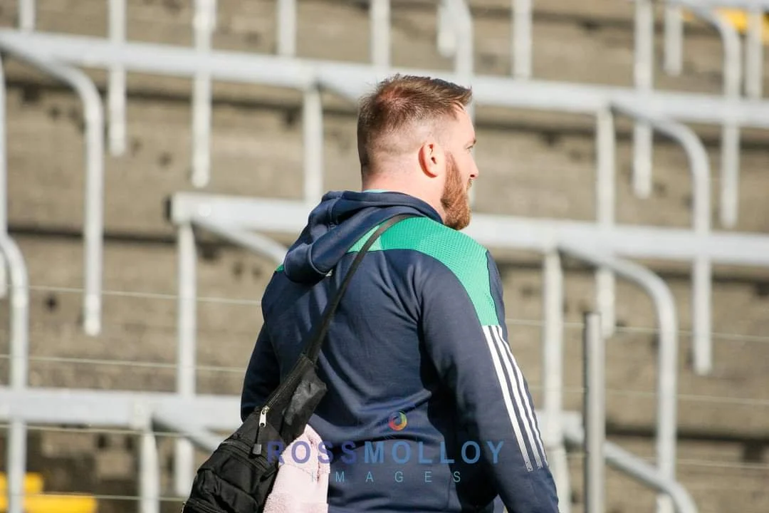A man with short red hair and a beard, wearing a navy blue hoodie with green and white stripes, carrying a black shoulder bag, standing on a stadium seating area.