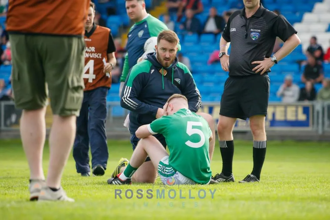 A soccer player in a green jersey sitting on the field clutching his head while a person in a green and navy jacket kneels beside him. A referee in black is standing nearby, and several people are in the background, some in orange jerseys.