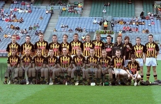 A soccer team in black and yellow striped jerseys and black shorts, posing for a group photo on a soccer field with empty stadium seats in the background.