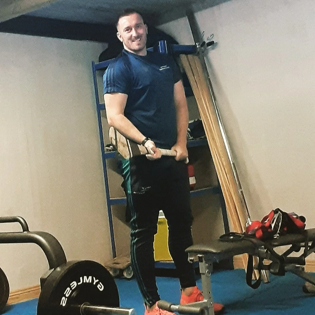 A man smiling in a gym holding a barbell with weights. He is wearing a navy T-shirt, black athletic pants with teal stripes, and salmon-colored sneakers. Equipment such as a weight bench, dumbbells, and wooden sticks are visible in the background.
