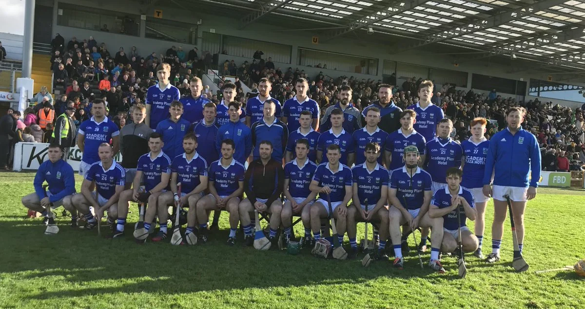 Group of hurlers in blue jerseys and shorts on a grass field, posing for a team photo with spectators in the background.